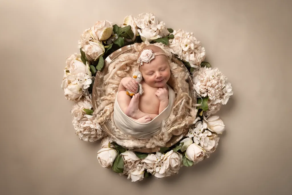 Nyfödd baby sover i rund skål omgiven av ljusa blommor och håller ett mjukisdjur.