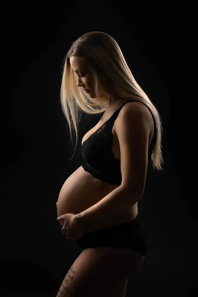 Gravid kvinna i profil håller om sin mage i svart studio med mjukt ljus.