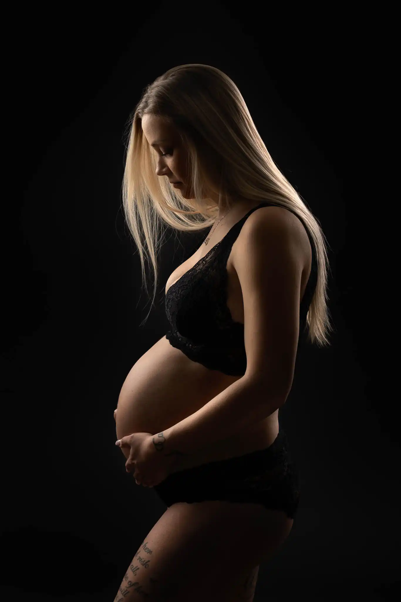 Gravid kvinna i profil håller om sin mage i svart studio med mjukt ljus.