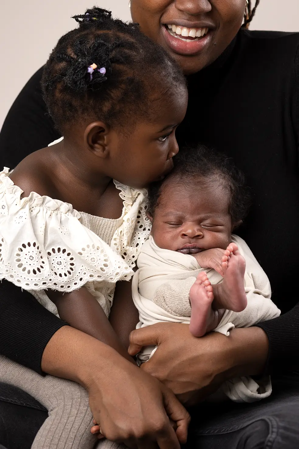 Kärleksfullt syskonmöte med bebis Syskon sitter i mammas knä, bebisen sover inlindad i filt.