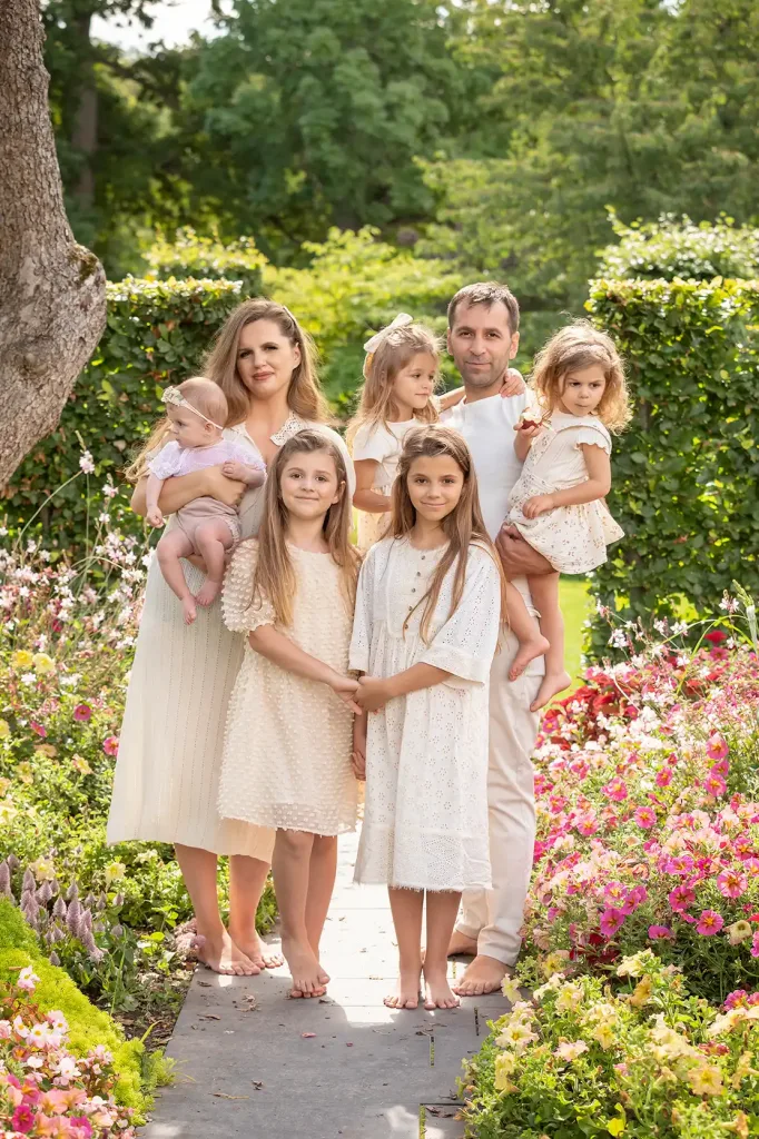 Familj med fem barn poserar barfota bland färgstarka blommor i trädgård.