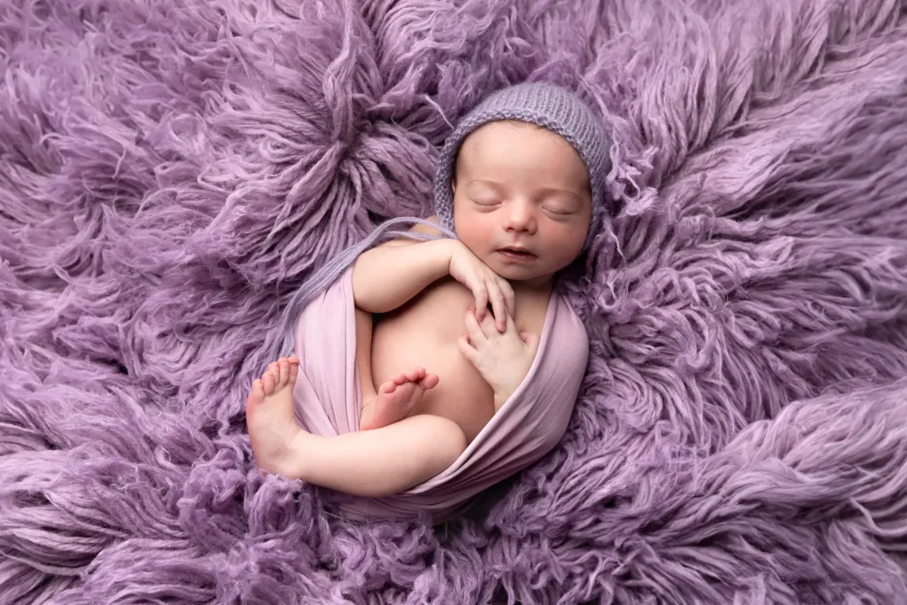 Sovande nyfödd baby inlindad i rosa wrap på fluffig lavendelfärgad filt.