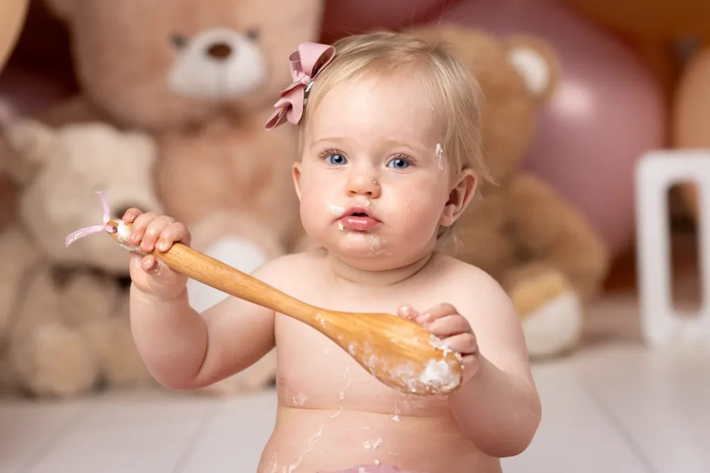 Halvnärbild på ettåring med träsked och tårta runt munnen under smash-the-cake-fotografering; suddiga nallar och ballonger i bakgrunden.