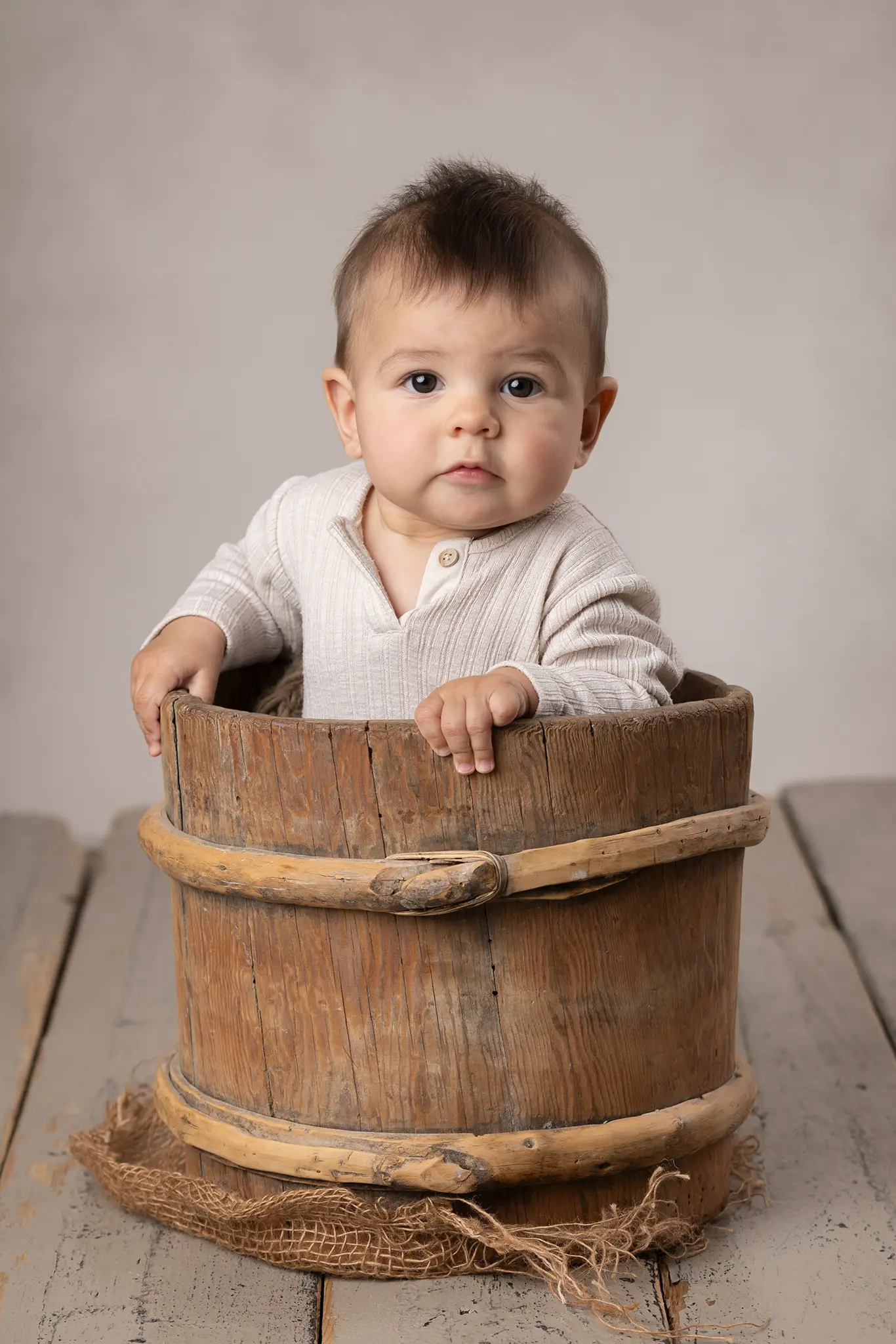 Bebis sitter i en trätunna och tittar mot kameran i studio med beige bakgrund.