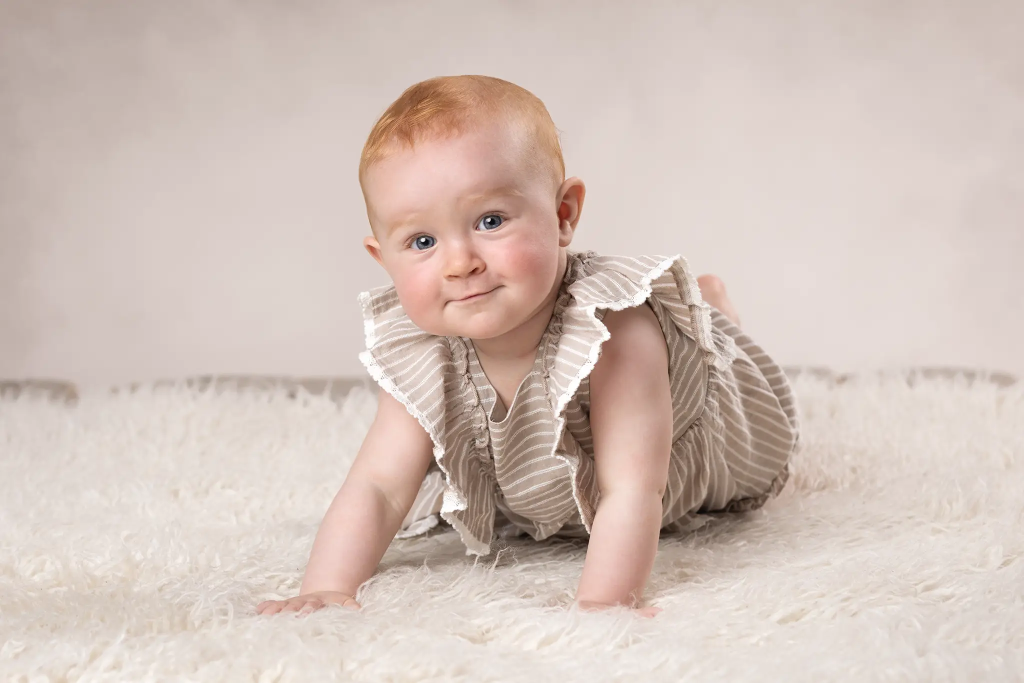 Bebis i beige klänning ligger på mage på ljus filt och tittar mot kameran.