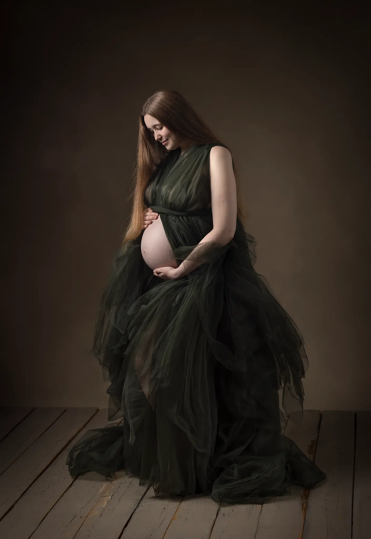 Gravidfotografering i mörkgrön klänning Gravid kvinna i mörkgrön tyllklänning står i studio och håller händerna om magen.