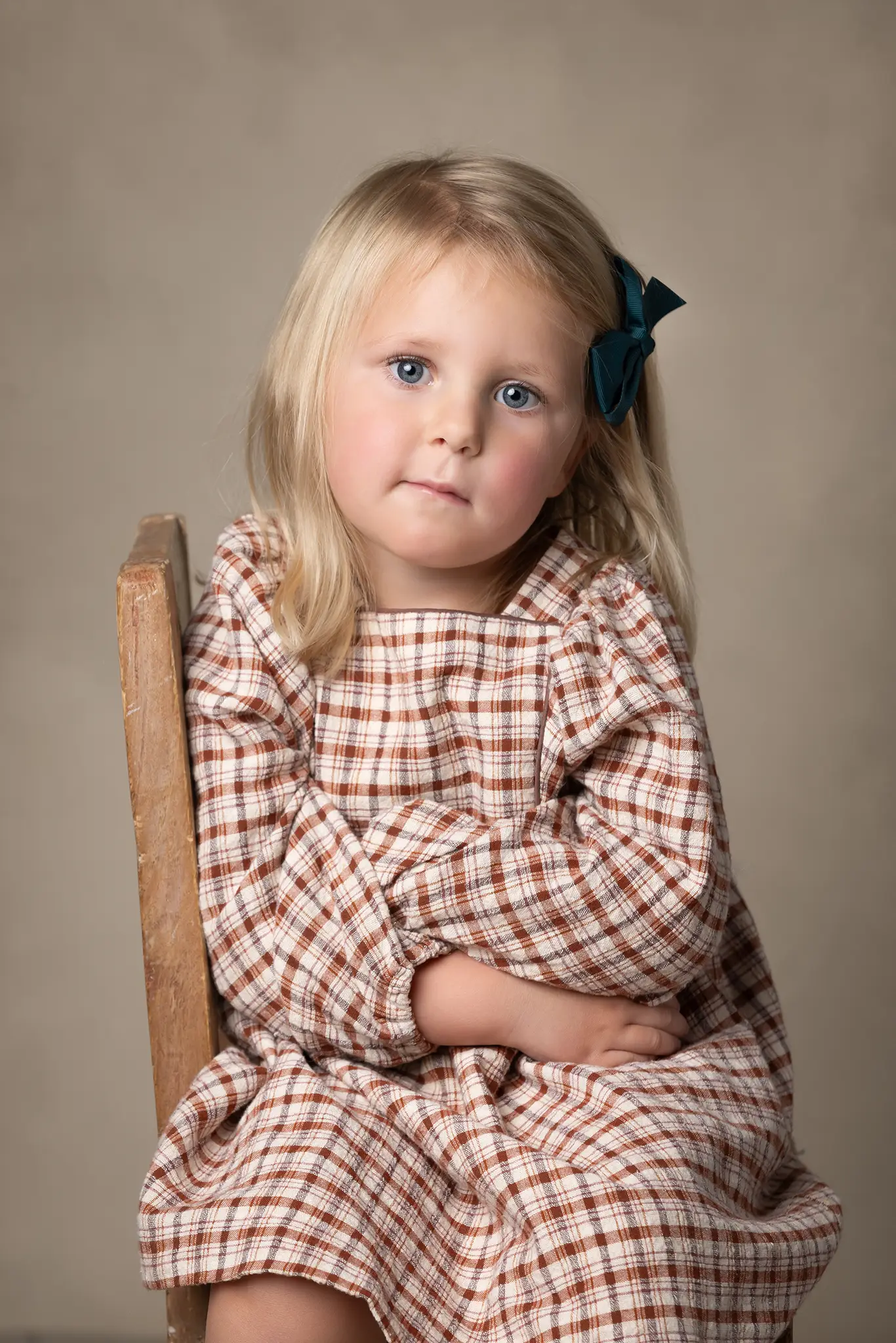 barnfotografering-lund-flicka-pa-trastol-rutig-klanning Barn sitter på trästol i studio mot neutral beige bakgrund, klädd i brunrutig klänning med mörkgrön rosett i håret.