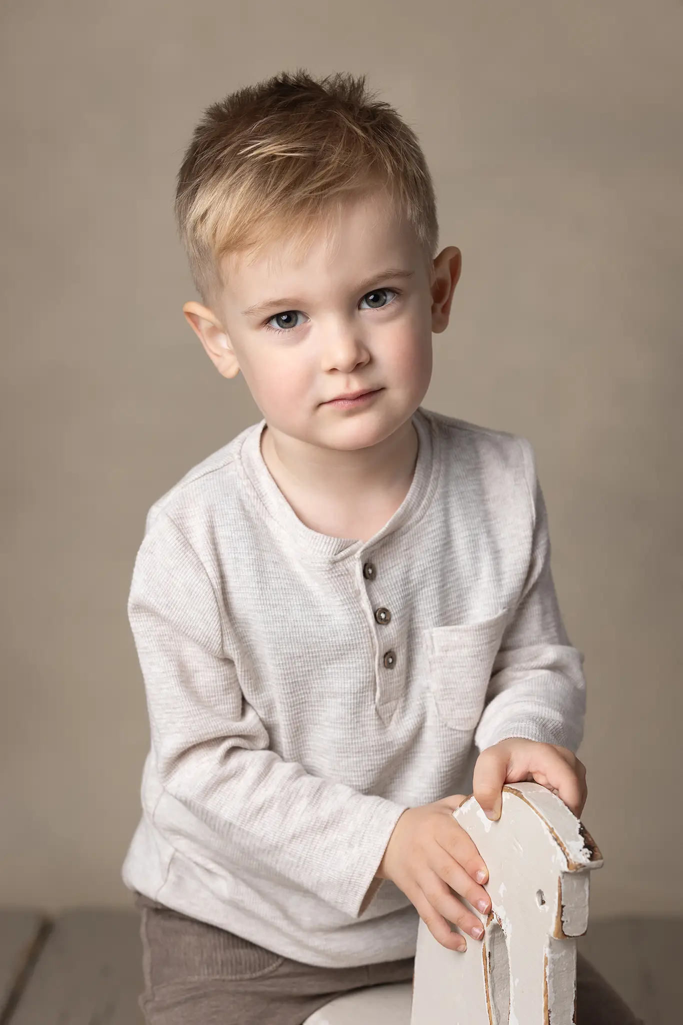 barnfotografering-lund-portratt-barn-vit-trahast Barn sitter i studio mot neutral beige bakgrund, tittar rakt in i kameran och håller i en vit trähäst.