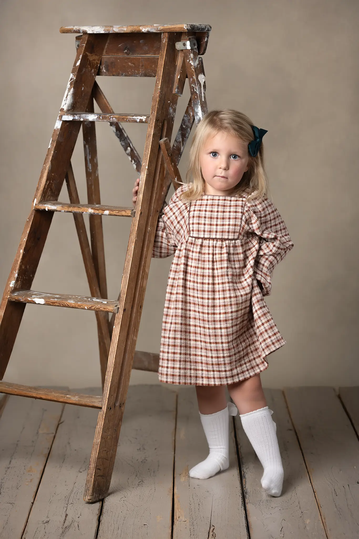Barnfotografering i klassisk stil Flicka i rutig klänning står bredvid en gammal trappstege i studio.