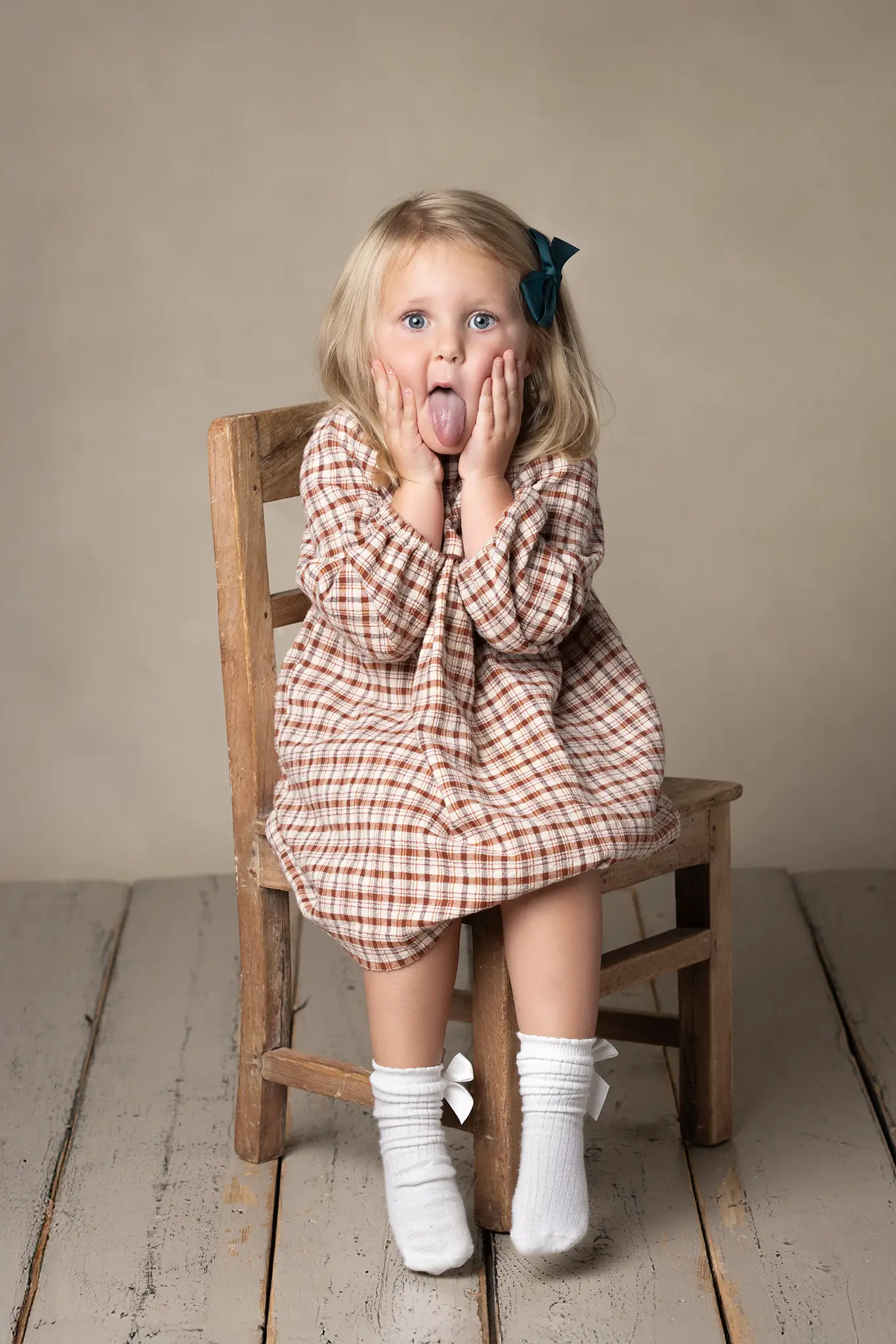 Lekfull barnfotografering i studio Flicka i rutig klänning sitter på en stol och gör en rolig min mot kameran.