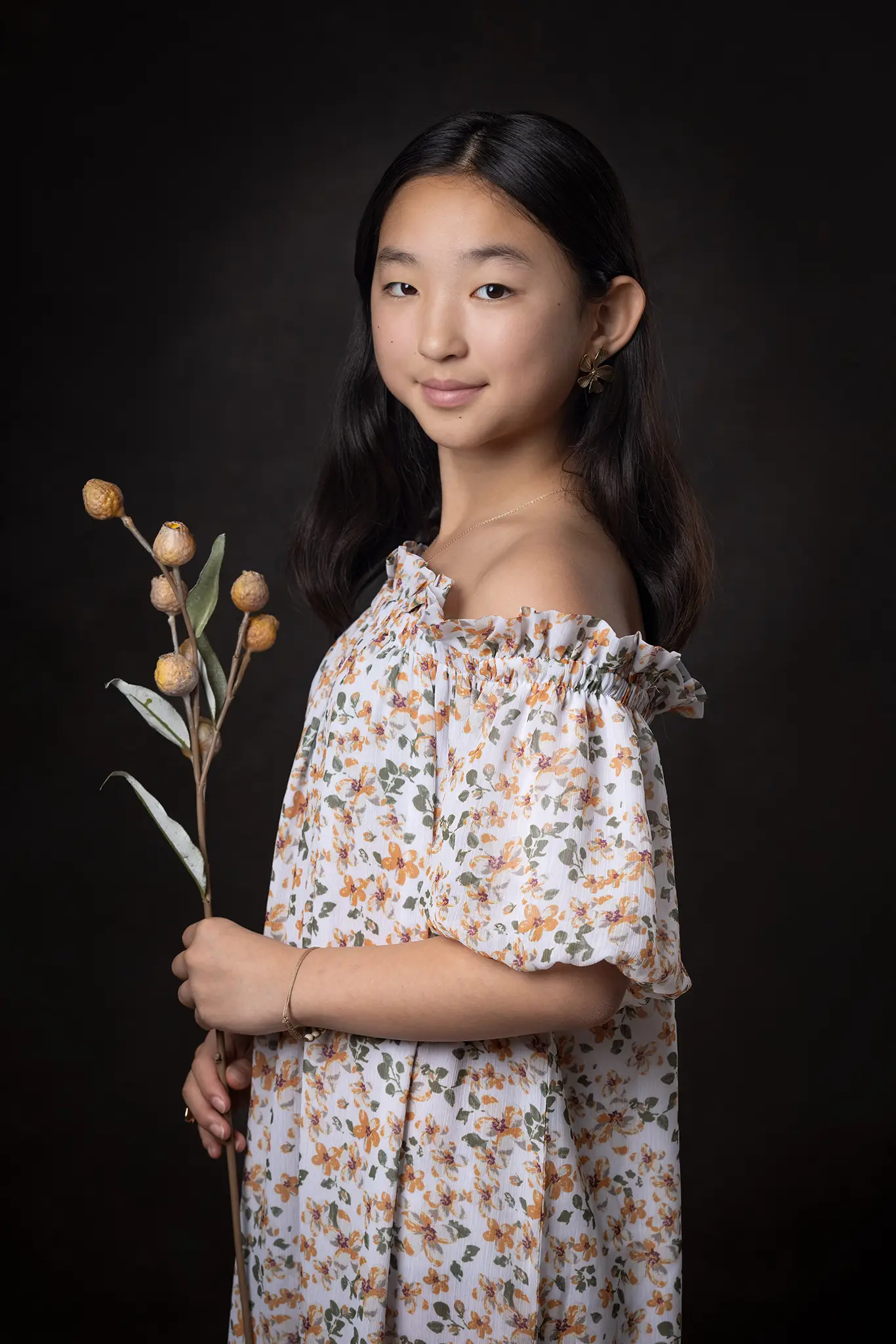 Ung flicka i blommig klänning håller en dekorativ kvist mot mörk studiobakgrund, klassiskt porträtt.