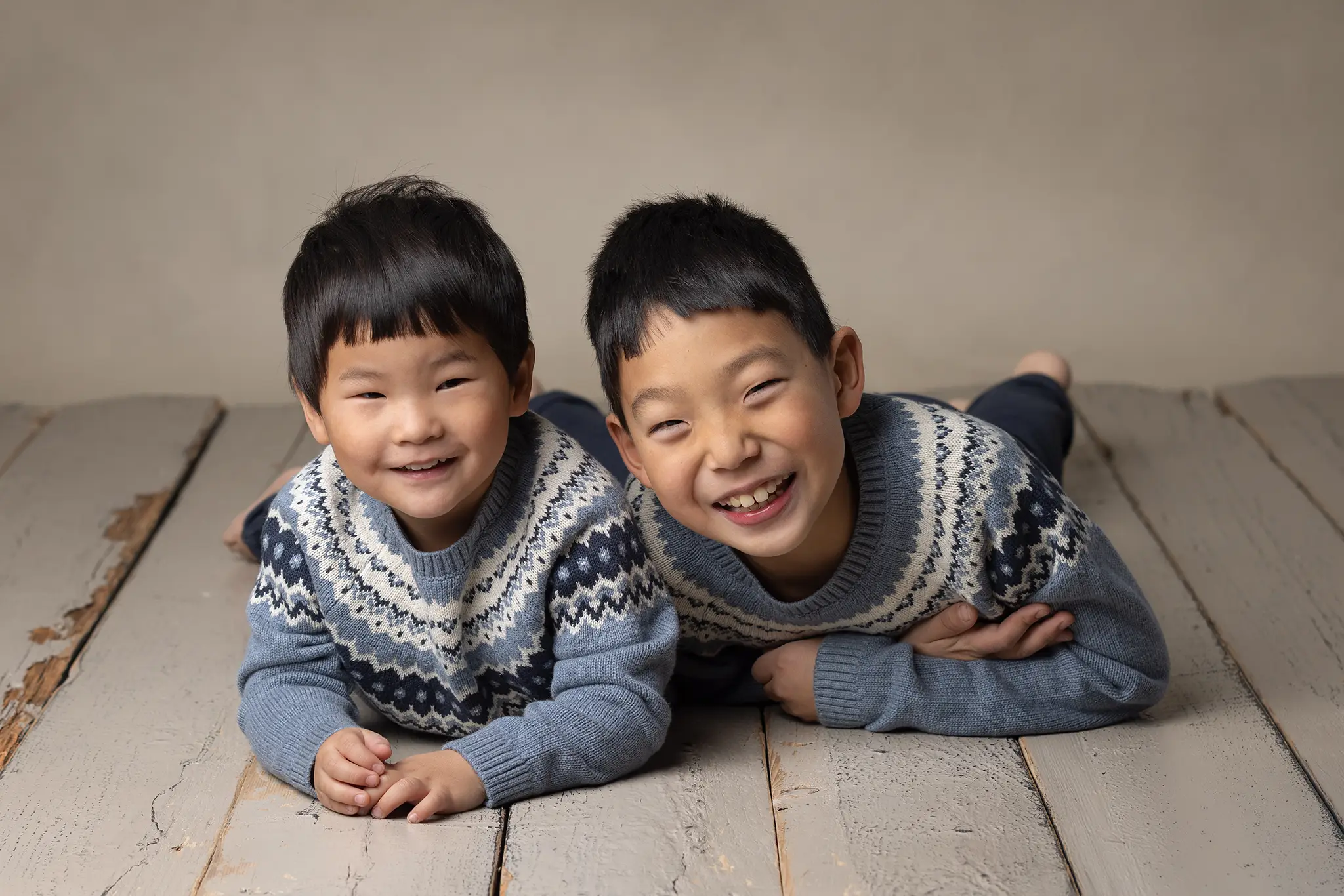 syskonfotografering-lund-tvabarn-ligger-pa-golv Två syskon ligger på studiogolv i matchande blå stickade tröjor och ler rakt in i kameran.