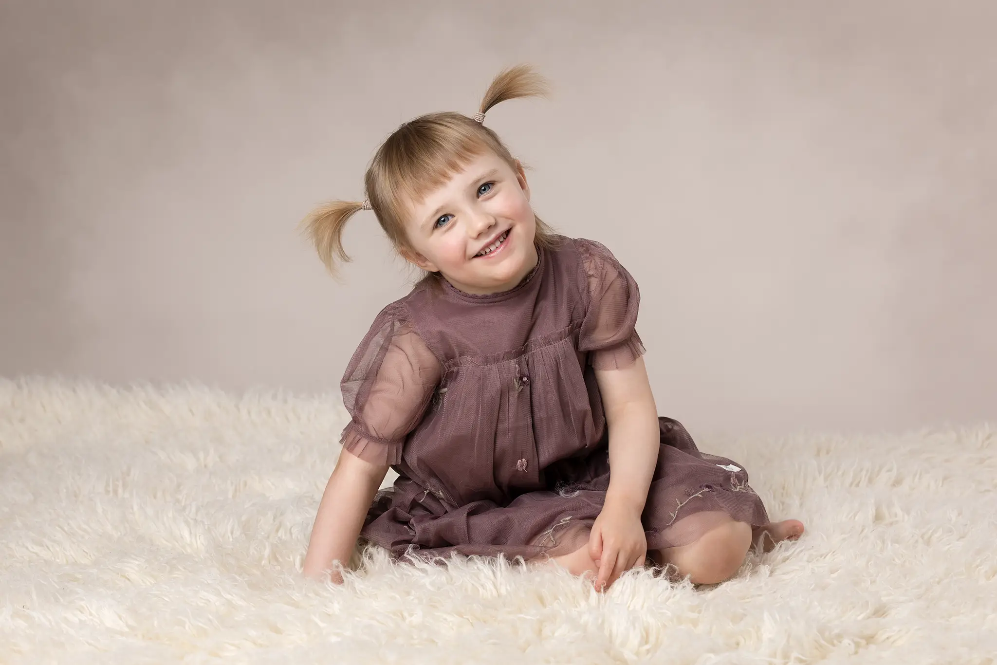 barnfotografering-lund-leende-flicka-pa-vit-filt Leende barn sitter på vit fluffig filt i studio, klädd i lila klänning, barnfotografering i Lund.