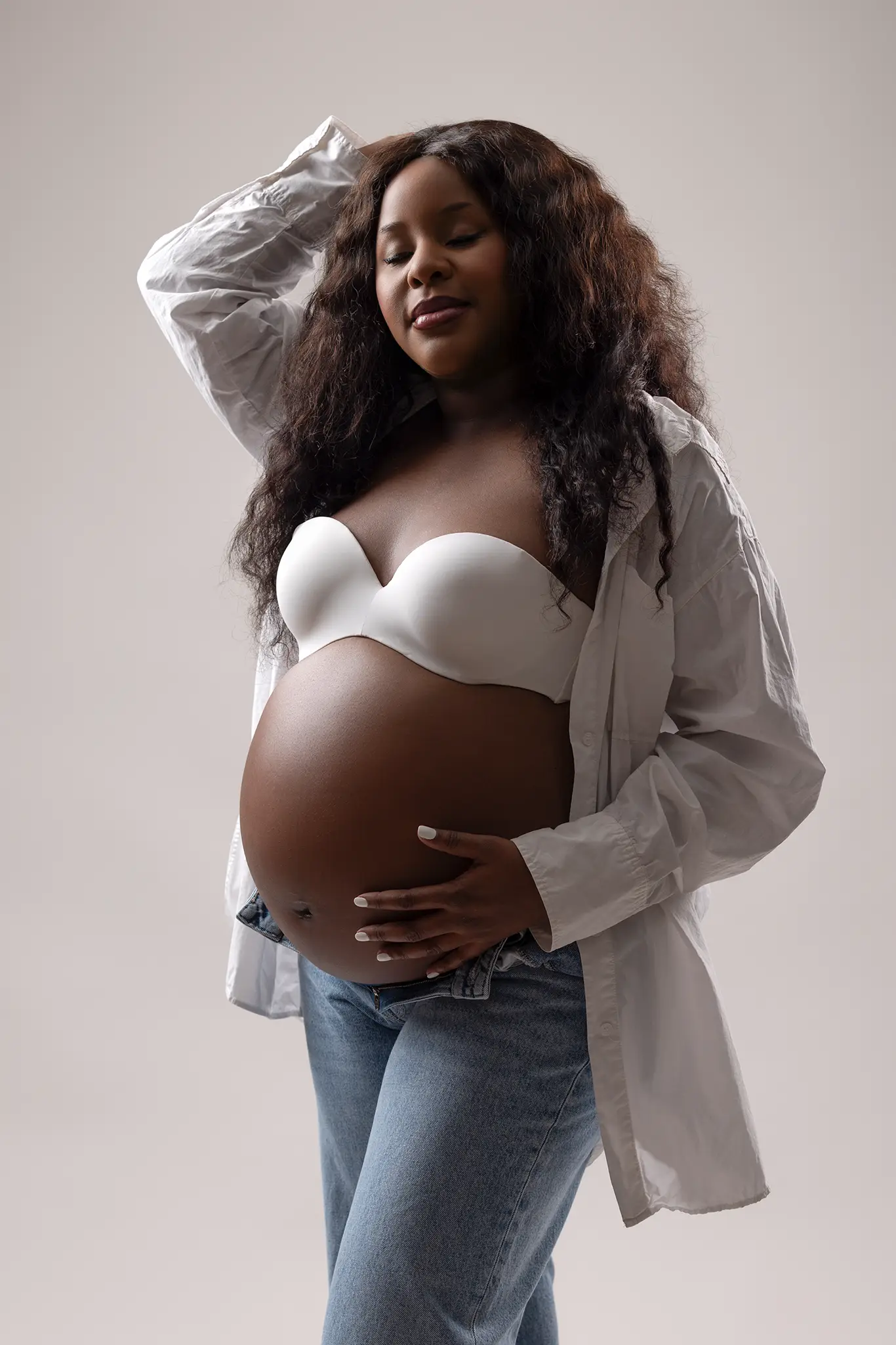 gravidfotografering-lund-gravidkvinna-vit-skjorta-jeans Gravid kvinna i vit axelbandslös bh och öppen skjorta håller om magen och blundar mot ljus, neutral studiobakgrund – gravidfotografering i Lund.