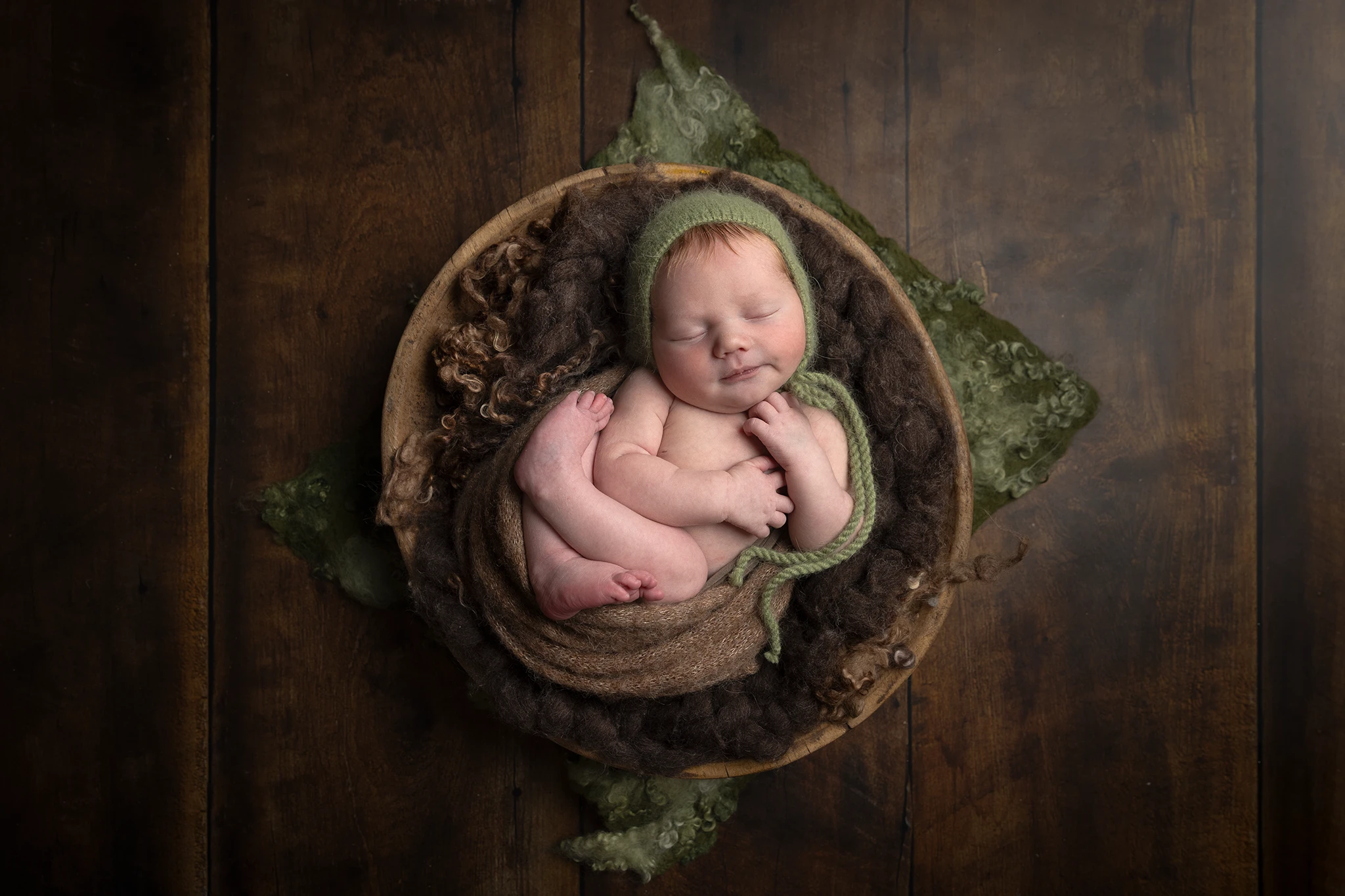 Nyfödd bebis sover hopkurad i träskål med brun ull och grön mössa, fotograferad uppifrån på trägolv – nyföddfotografering i Lund.