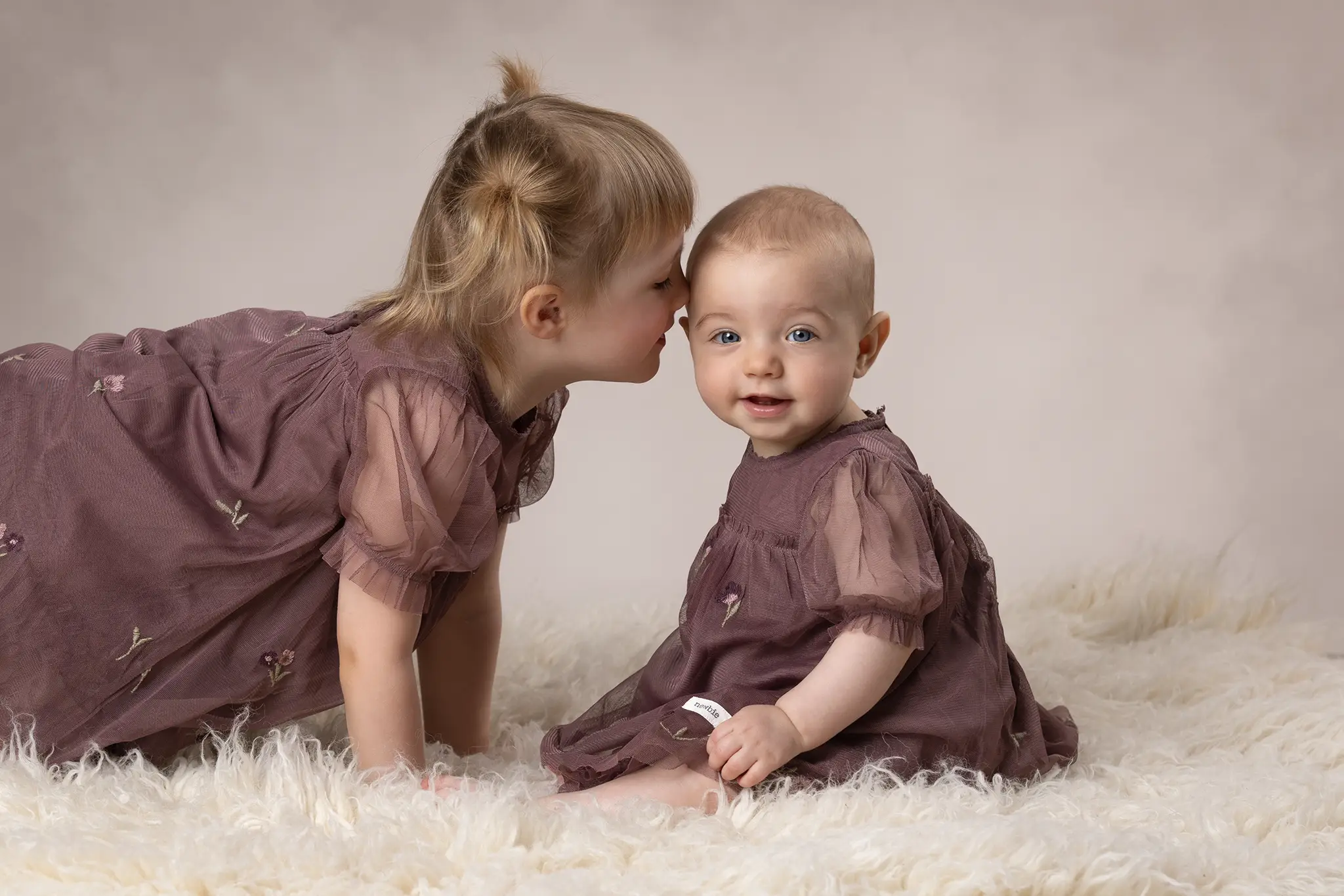 Äldre syskon lutar sig fram och viskar i örat på sitt yngre syskon som sitter och ler mot kameran på en vit, fluffig filt i studio.