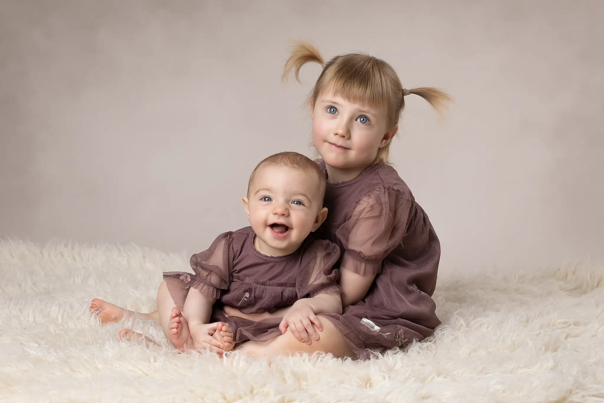 syskonfotografering-lund-storasyster-haller-bebis-pa-vit-filt Två små syskon i matchande lila klänningar sitter på vit fluffig filt i studio, bebisen ler mot kameran i famnen på storasyster.