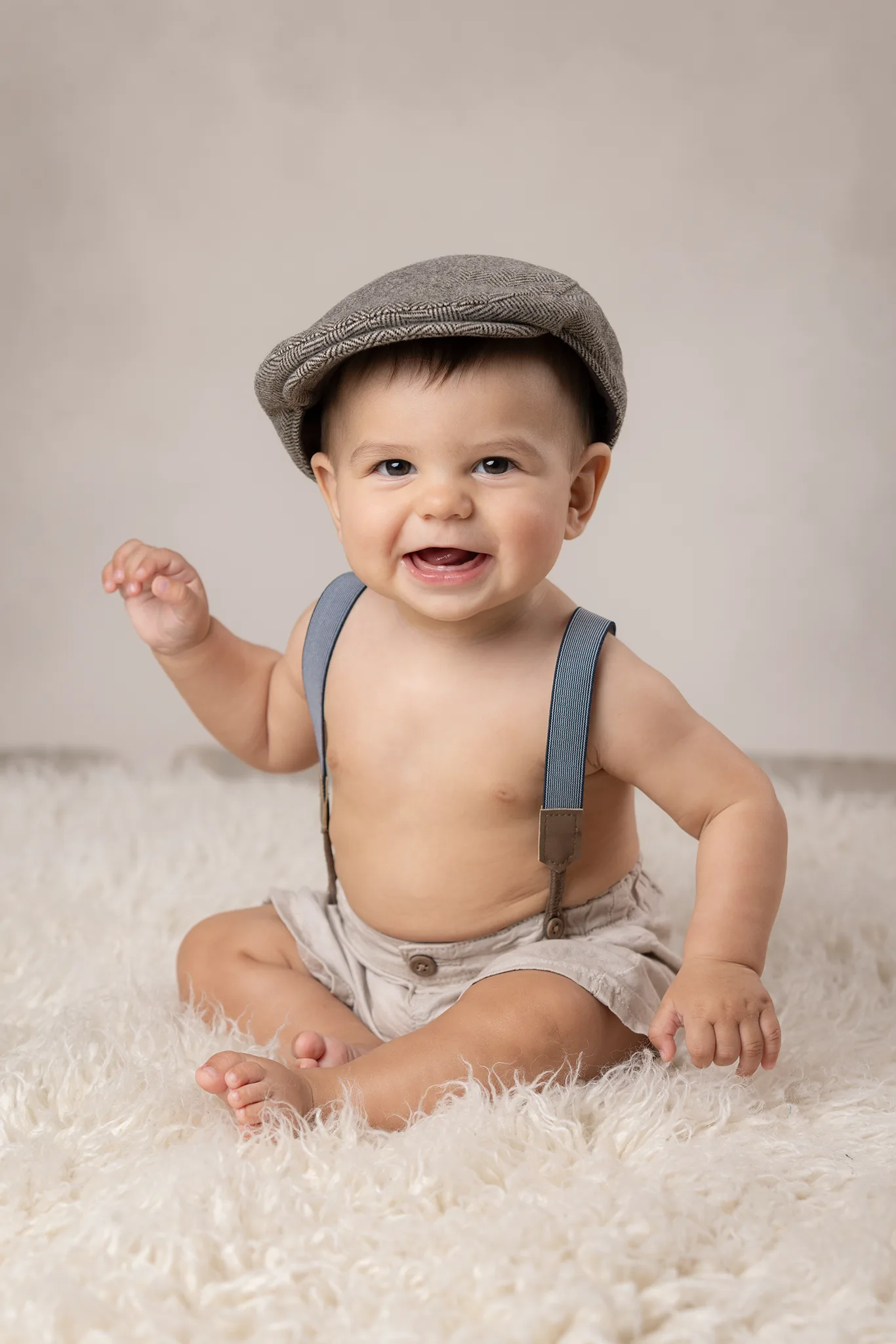 Bebis sitter på ljus fäll mot beige bakgrund med keps och hängslen