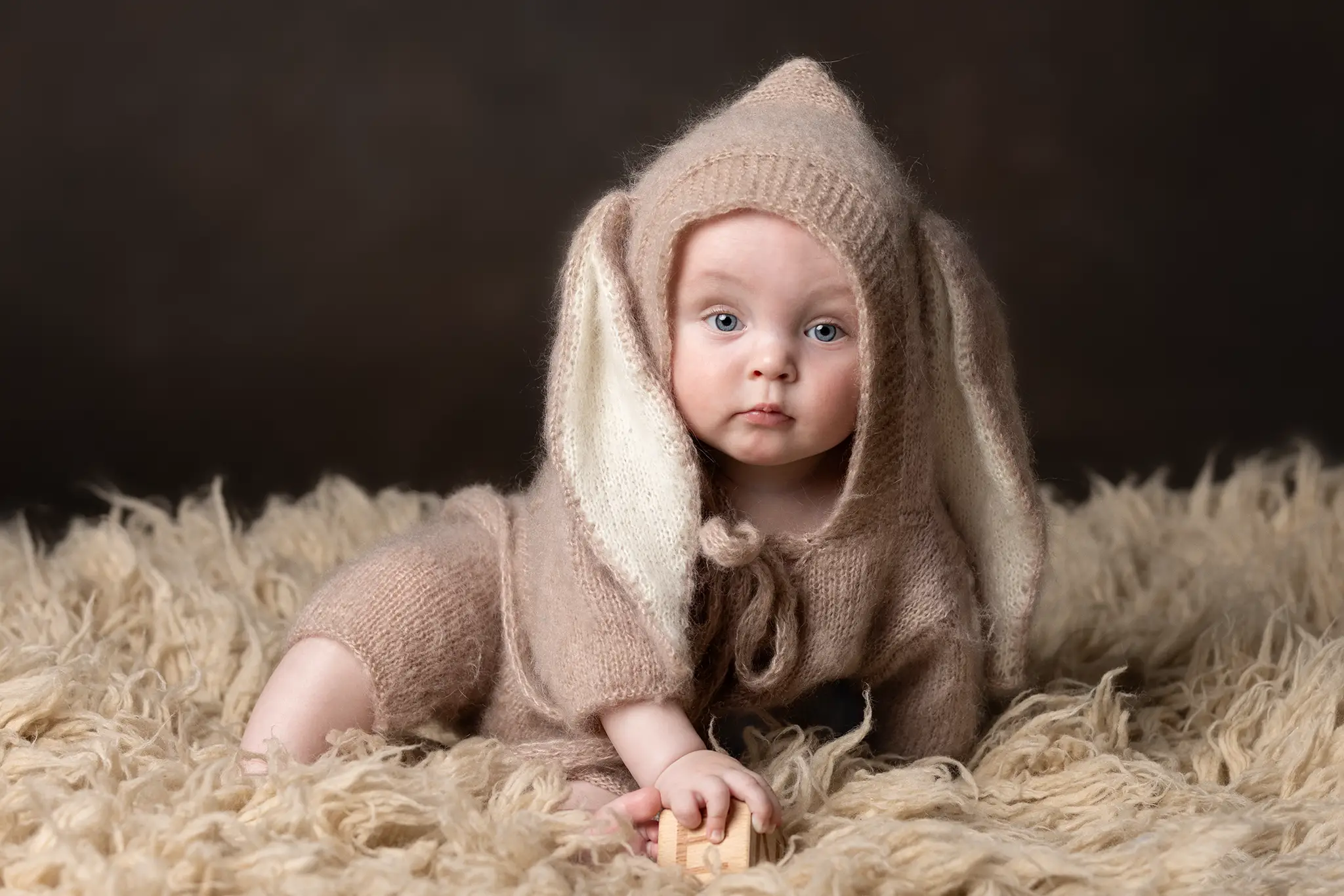 barnfotografering-kanindrakt-pasktema Bebis i stickad kaninhuva på lurvig filt i studio, håller en liten träkloss och ser in i kameran.