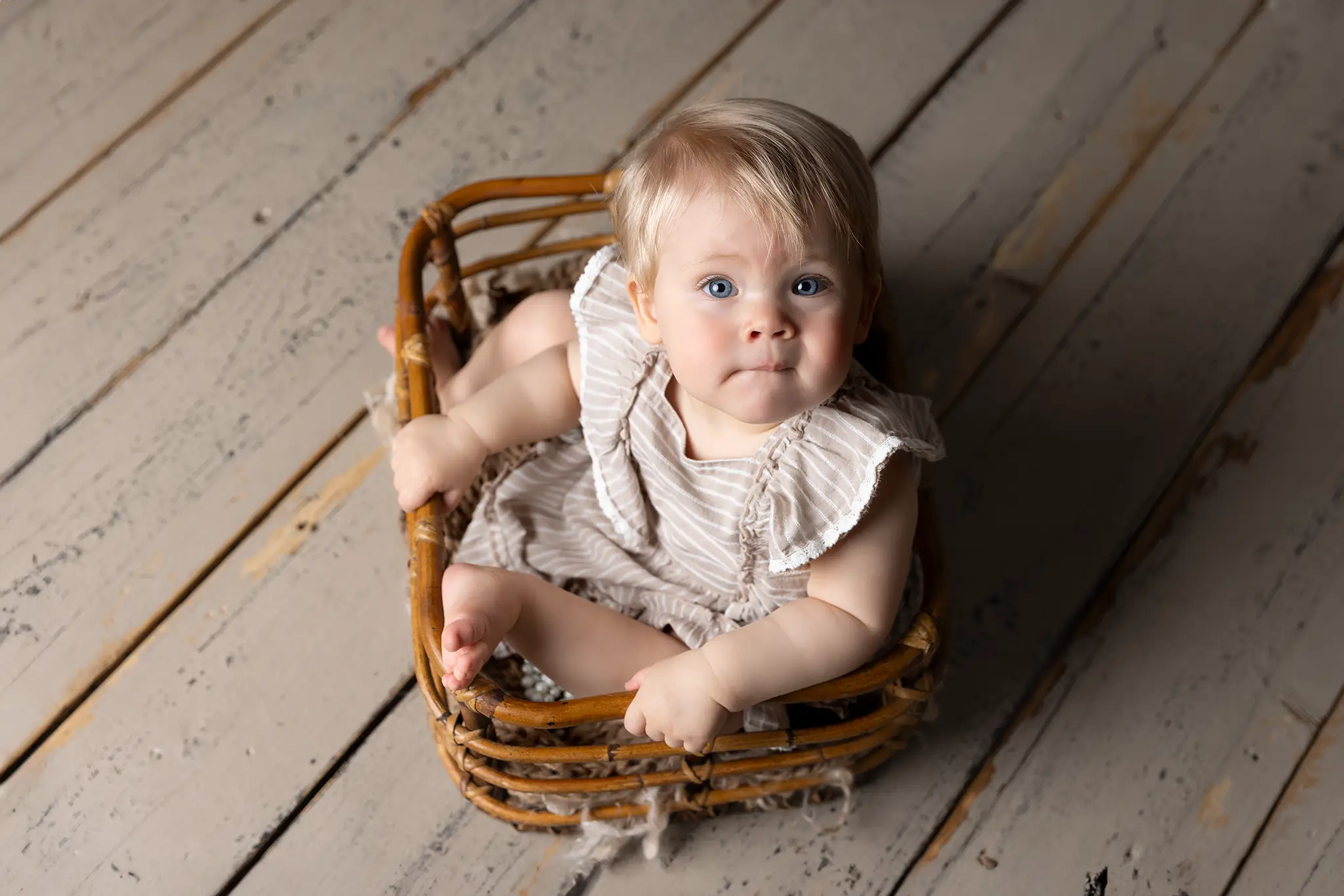 bebis-rottingkorg-portratt-studio En 1-årig flicka sitter i en korg på trägolv.