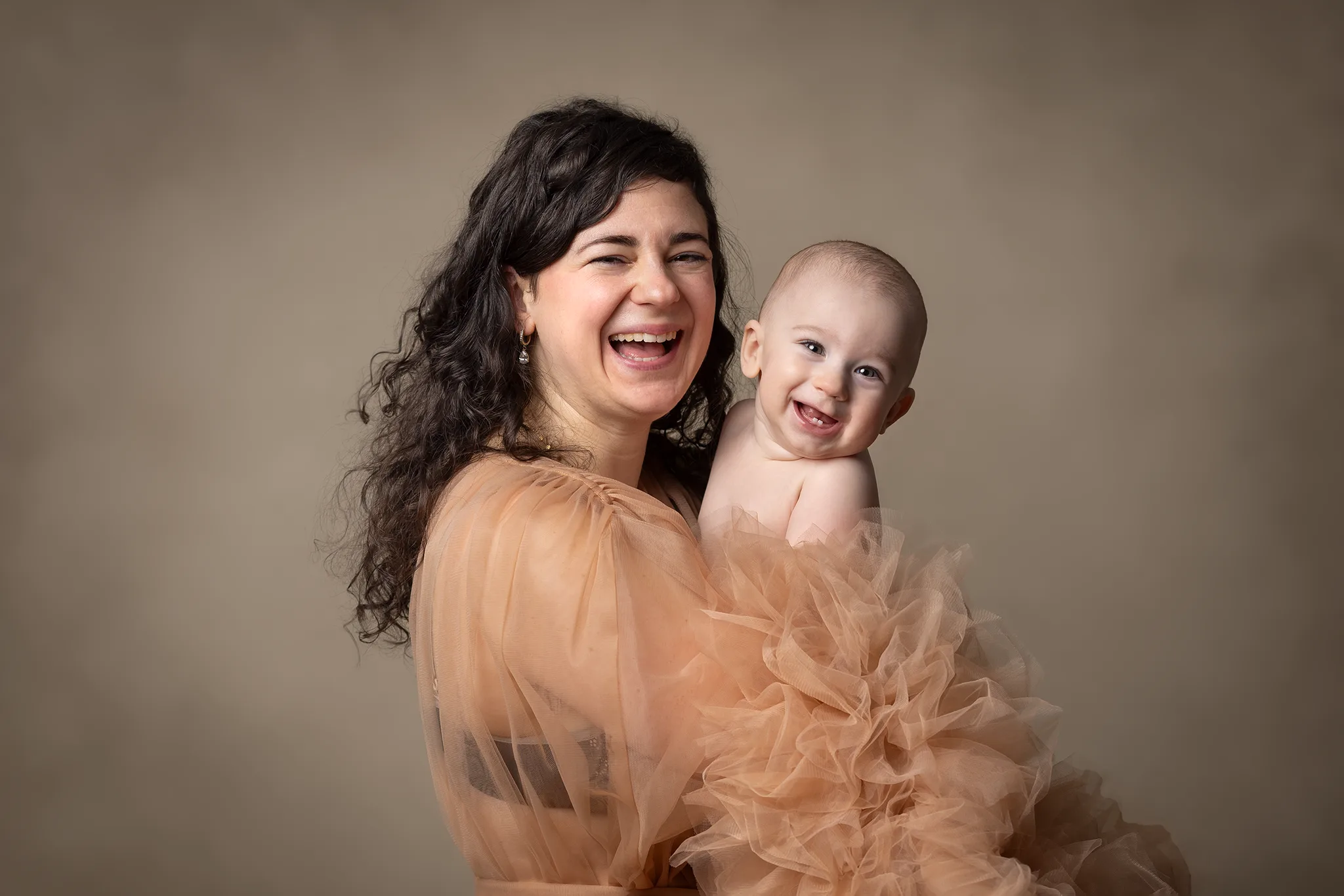 Leende förälder håller glad bebis i studio, båda i varma beige toner.