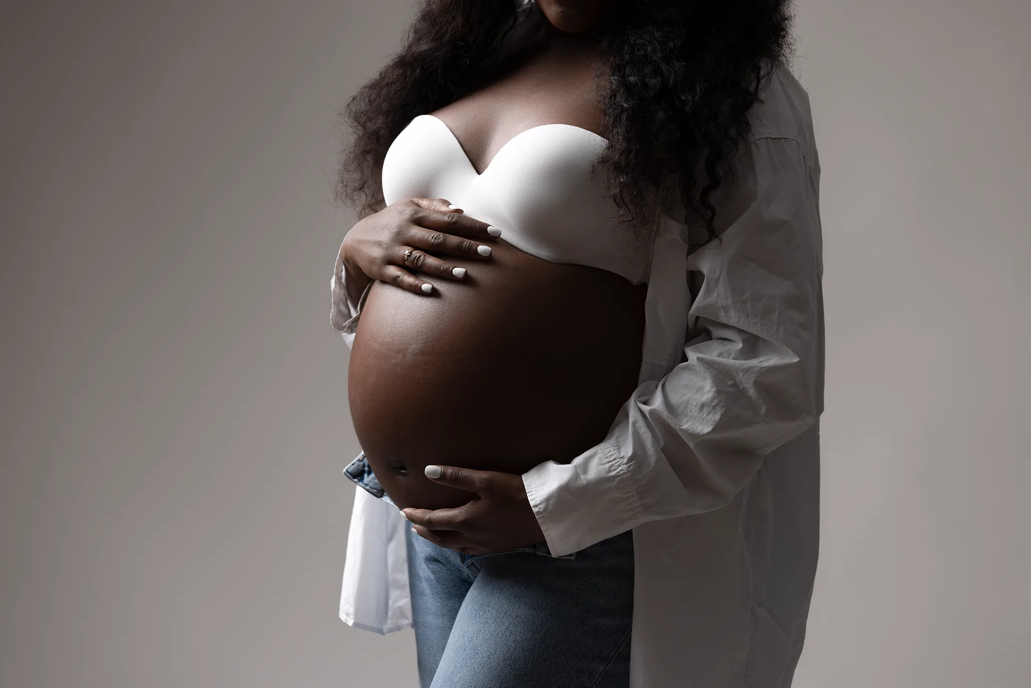 gravidfotografering-detalj-gravidmage-vit-bh-skjorta-jeans Närbild av gravid kvinna i vit bh, skjorta och jeans som håller händerna runt sin nakna gravidmage i studio.