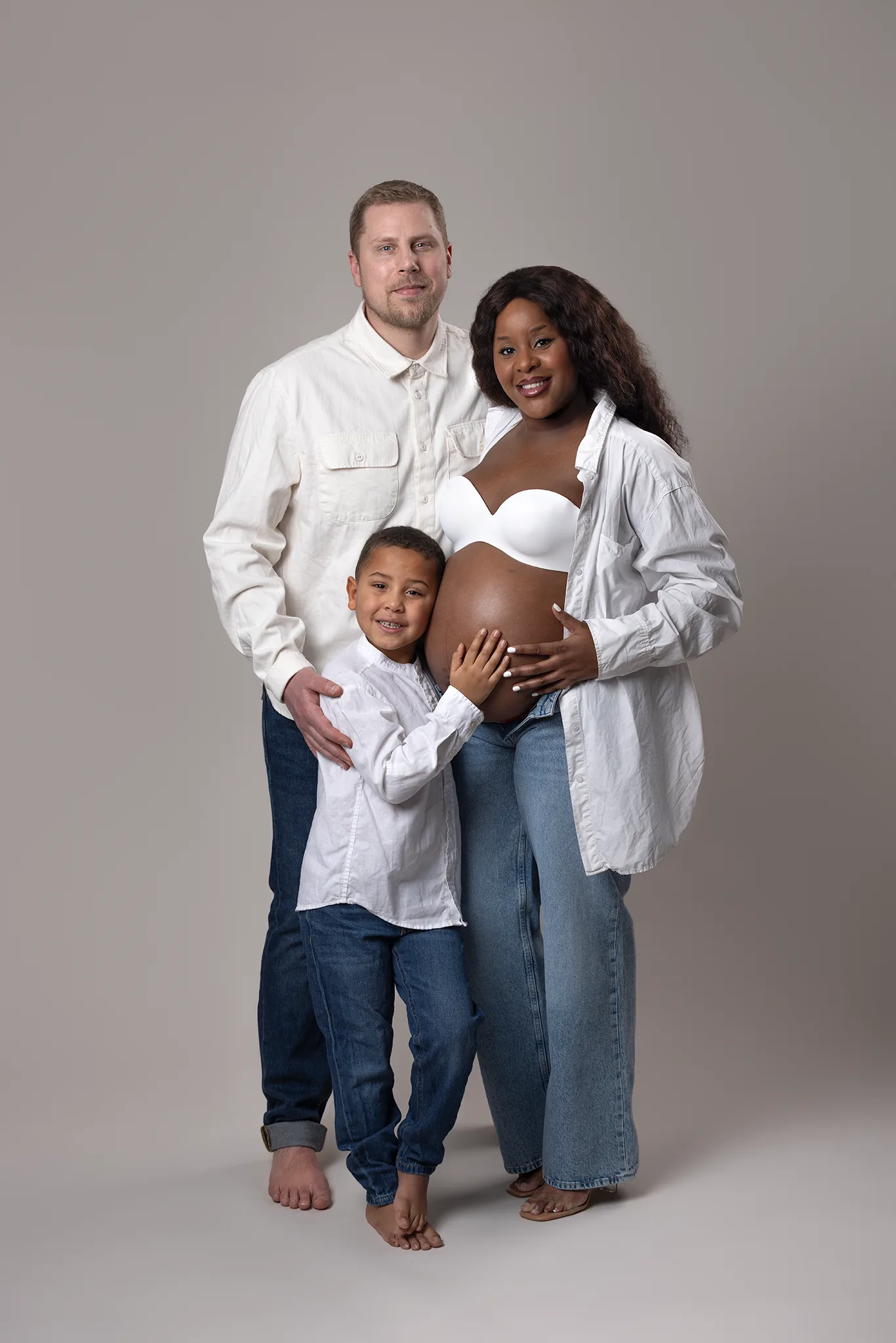 gravidfotografering-lund-familj-med-barn-hander-pa-gravidmage Gravid familj i studio där föräldrarna står tillsammans med sitt barn som håller händerna på mammans gravida mage mot ljus, neutral bakgrund.