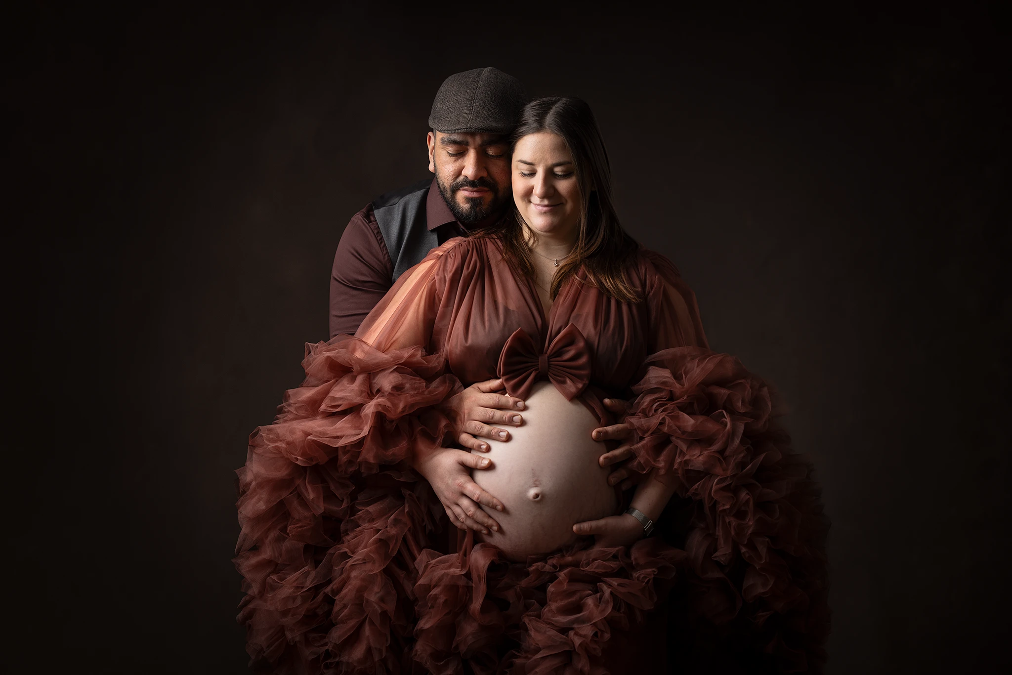 gravidfotografering-lund-par-haller-om-gravidmage-rustbrun-tyll Gravid kvinna i rustbrun tyllklänning står mot mörk bakgrund med partnern bakom sig, båda håller händerna runt gravidmagen.