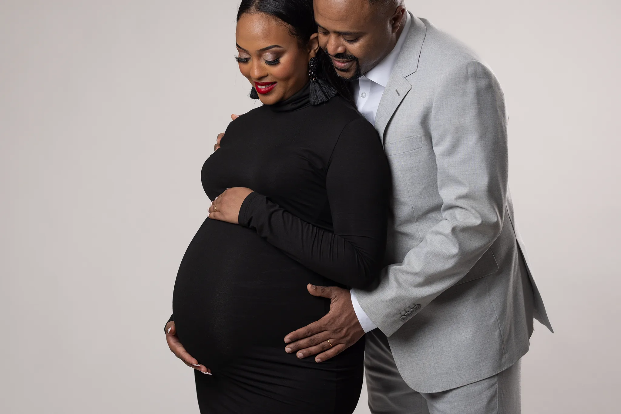 gravidfotografering-lund-par-haller-om-gravidmage-studio Gravid kvinna i svart klänning står i studio med partner i ljusgrå kostym bakom sig, båda håller händerna runt gravidmagen.