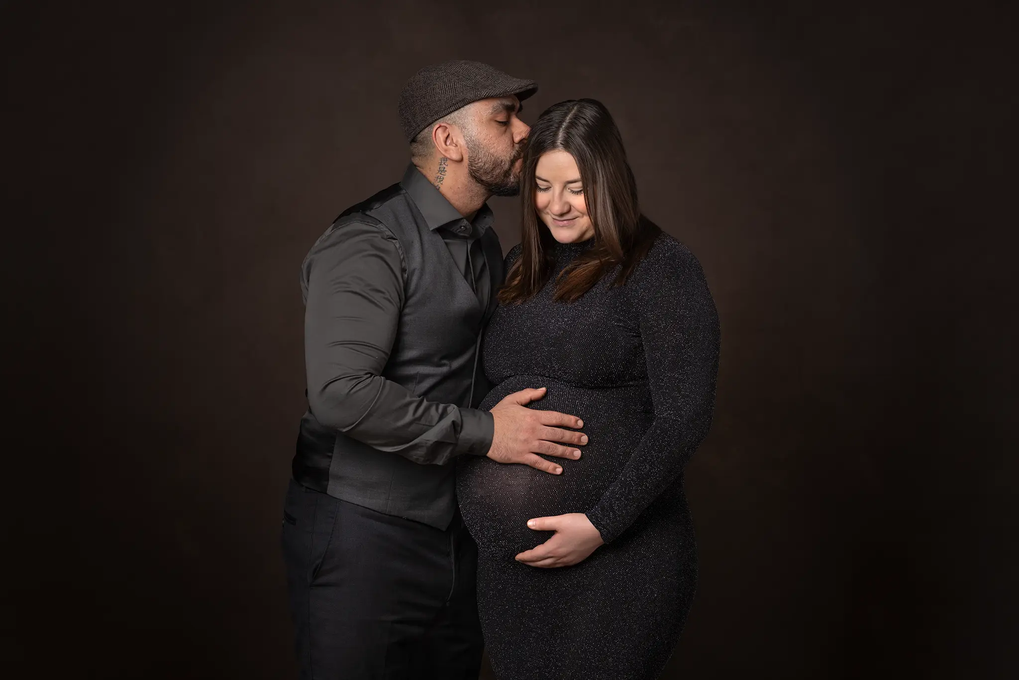 gravidfotografering-lund-par-kysser-panna-mork-bakgrund Gravid kvinna i glittrig svart klänning står mot mörk studiobakgrund medan partnern kysser hennes panna och håller händerna på magen.