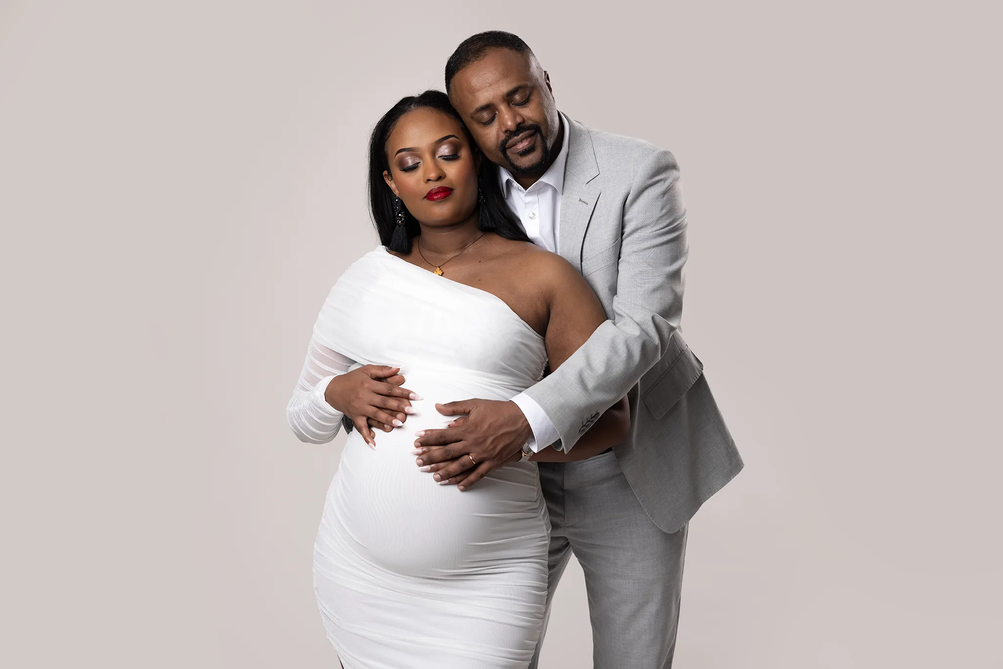 gravidfotografering-lund-par-vit-klanning-gra-kostym-hander-pa-magen Gravidfotografering i vår studio i Lund där paret står tätt tillsammans mot en ljus, neutral bakgrund. Den blivande mamman bär en vit klänning och partnern i grå kostym håller om henne med händerna över gravidmagen.