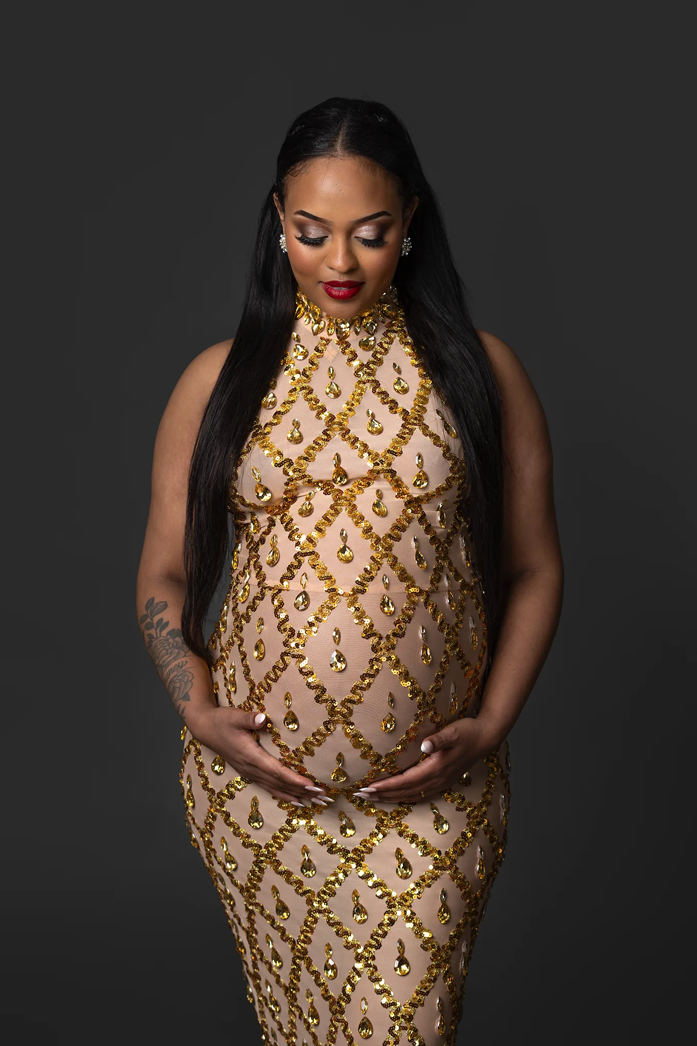 Gravid kvinna med rött läppstift och lång paljettklänning i guld står i studio i Lund och håller om sin mage.