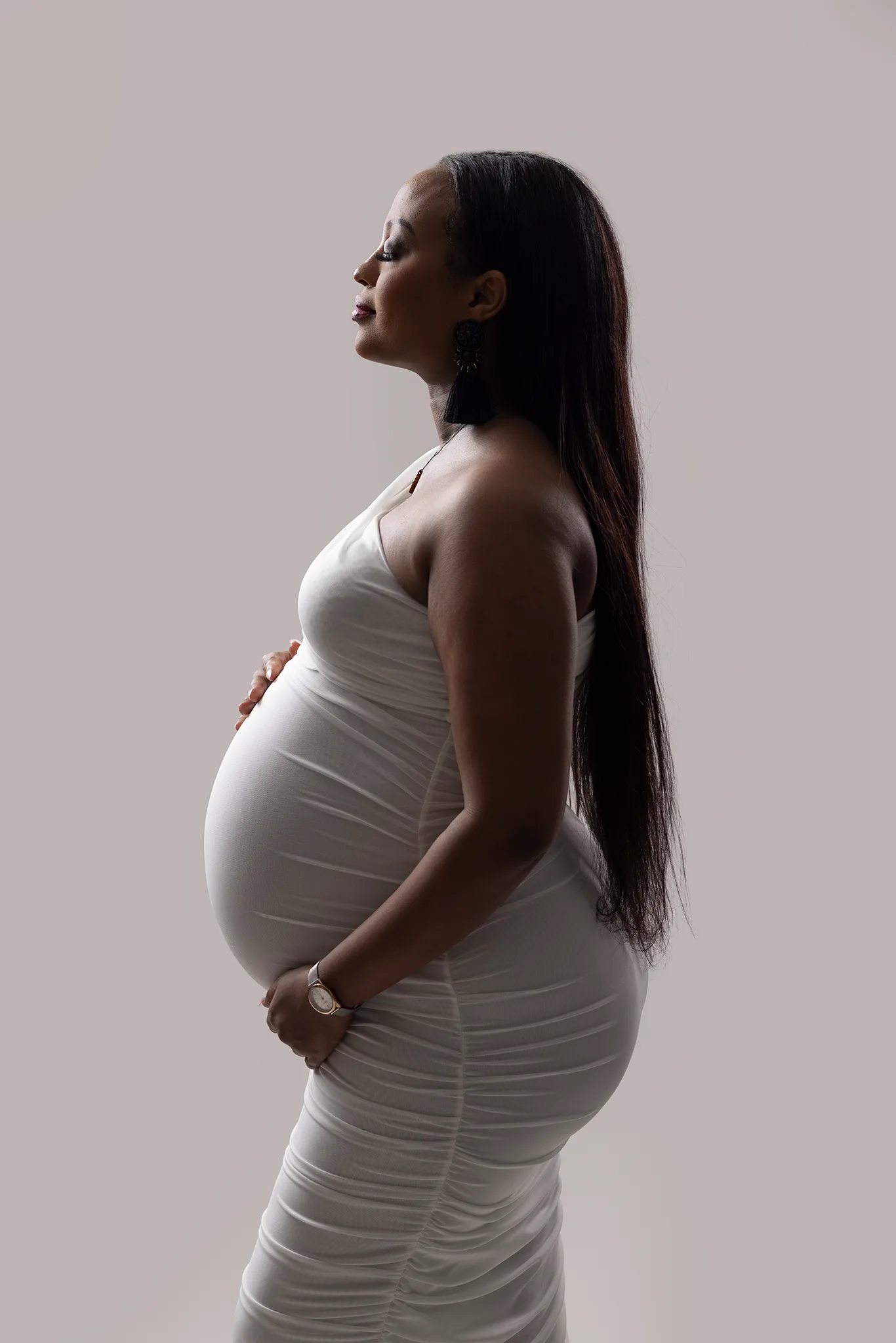 gravidfotografering-profilfoto-gravidkvinna-neutral-bakgrund Gravid kvinna i vit åtsittande klänning står i profil mot ljus, neutral bakgrund med händerna på magen.