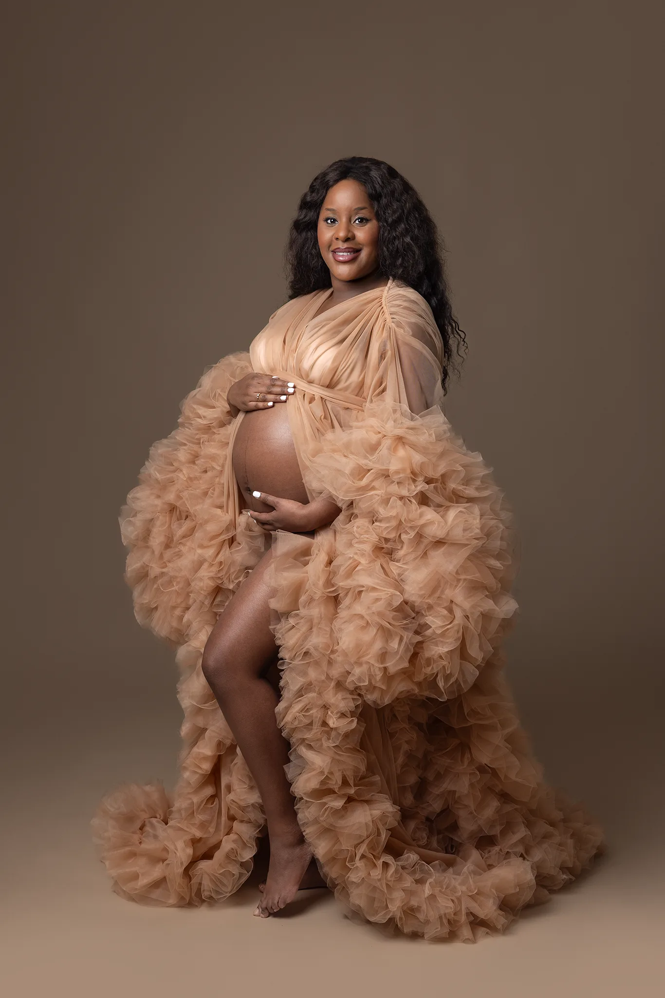 Gravid kvinna i aprikosfärgad tyllklänning står barfota mot mörk brun studiobakgrund och håller händerna runt sin nakna gravidmage.