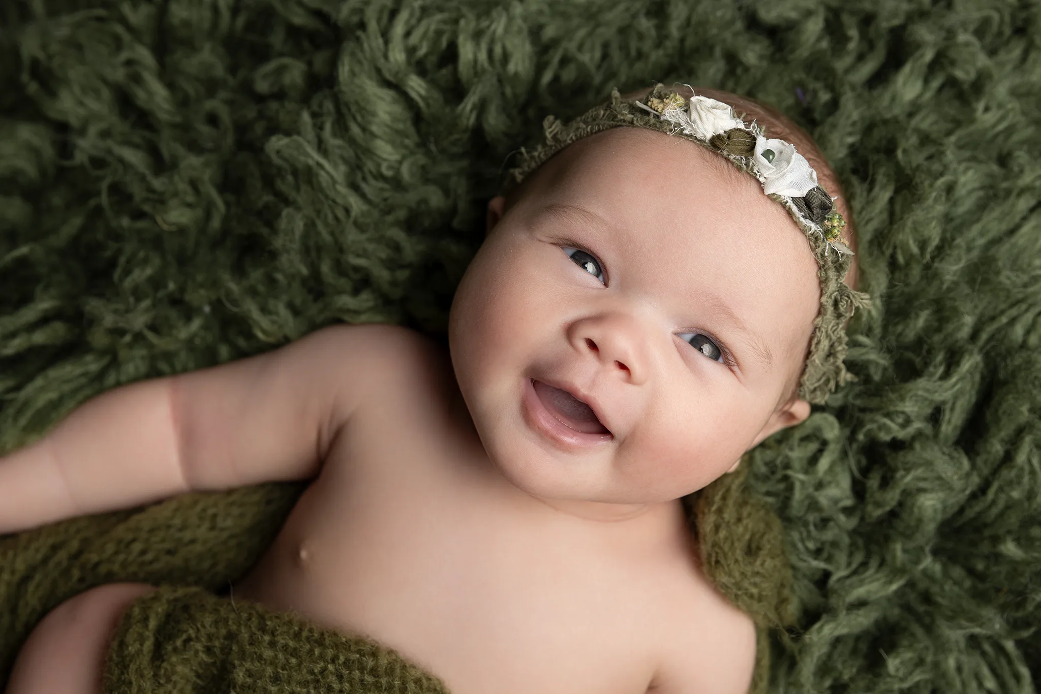 babyfotografering-lund-nyfodd-leende-gron-filt-blomsterband Närbild av leende nyfödd på grön lurvig filt, med blomsterband runt huvudet.