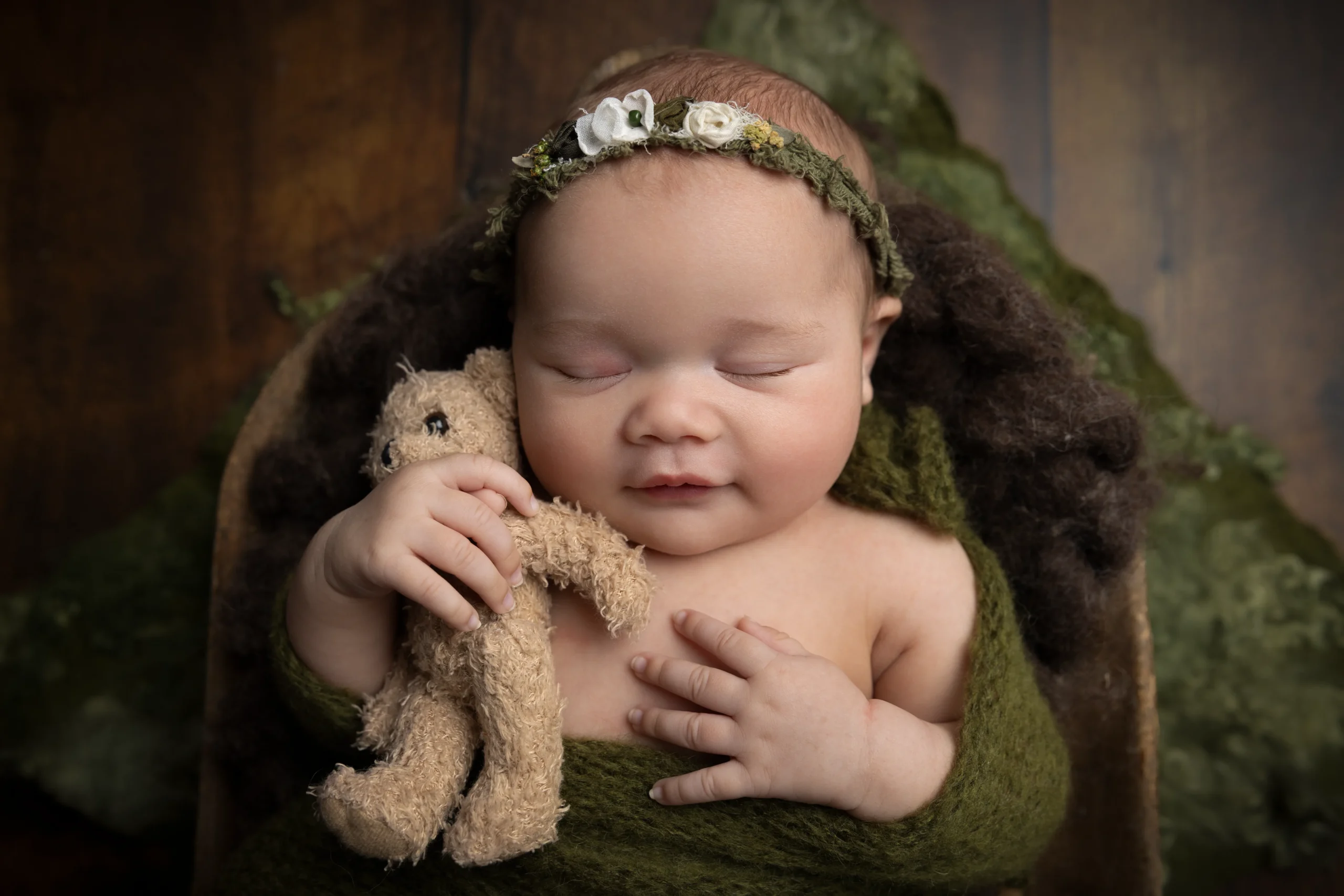 Sovande nyfödd med blomsterband håller en liten nalle, inlindad i grön wrap i studio.