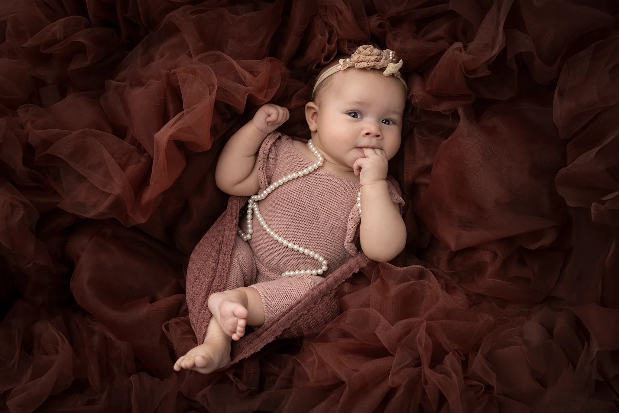 familjefotografering-lund-kreativt-babyportratt-tyll Närbild av baby med hårband och pärlhalsband, liggande i vinröd tyll.