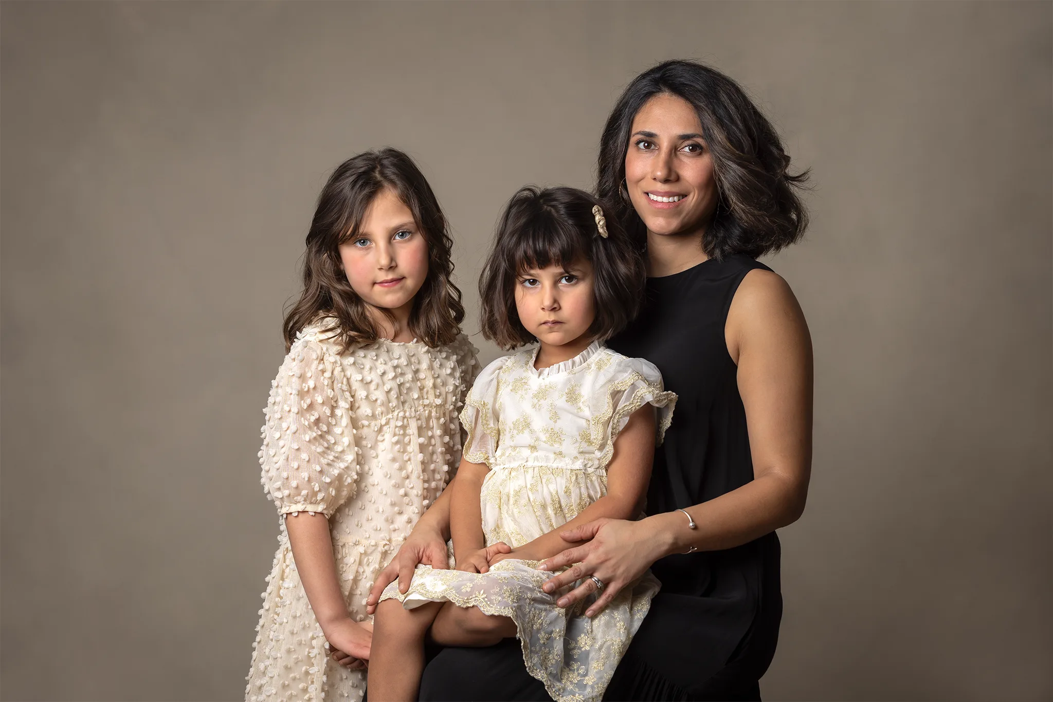 Studioportätt av en vuxen och två barn i ljusa klänningar mot neutral bakgrund.