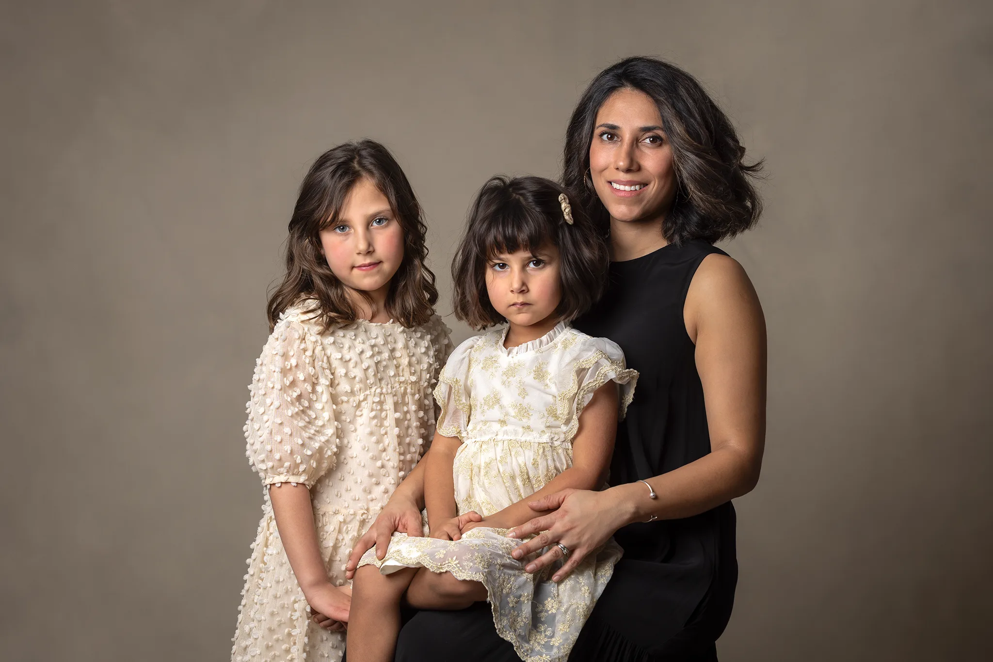 Studioportätt av en vuxen och två barn i ljusa klänningar mot neutral bakgrund.
