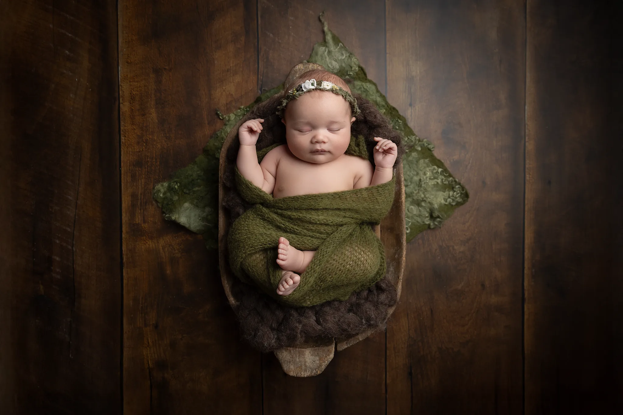 Nyfödd baby sover lindad i grön wrap i en skål på mörkt trägolv.