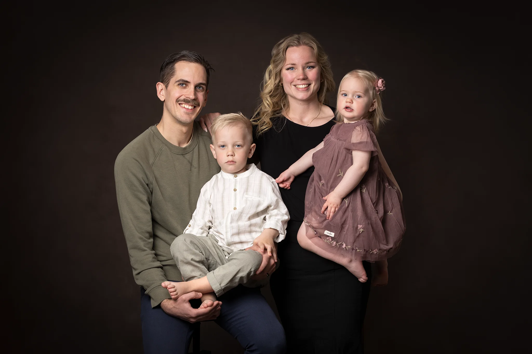 Familjeporträtt i studio: leende mamma och pappa håller sina två små barn, familjefotografering i Lund.