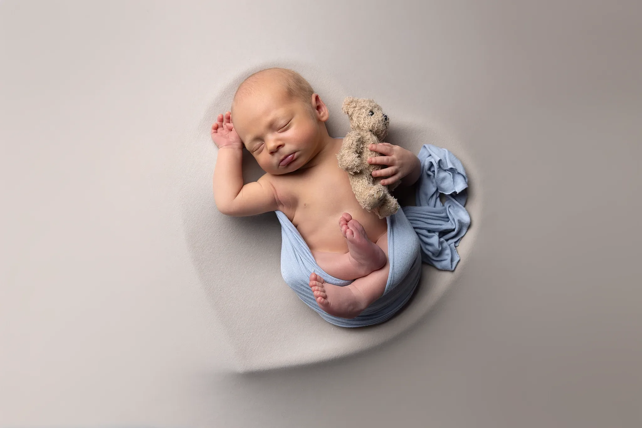 nyfoddfotografering-lund-bebis-bla-wrap-nalle Nyfödd pojke sover invirad i ljusblå sjal och kramar en beige nalle, fotograferad ovanifrån i Lund.