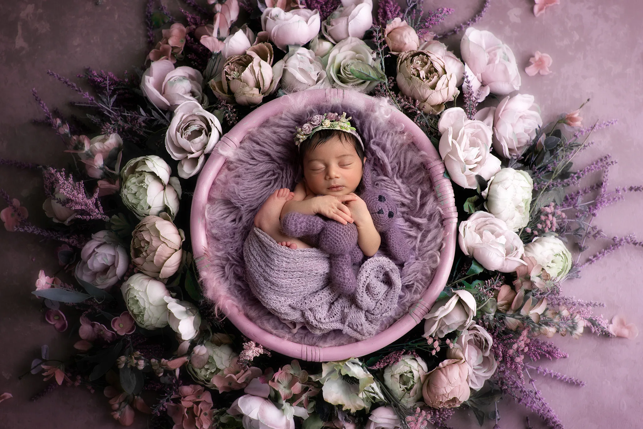 Nyfödd bebis sover hoprullad i lila skål omgiven av pastellrosor, håller en stickad kanin; studiofoto i Lund.