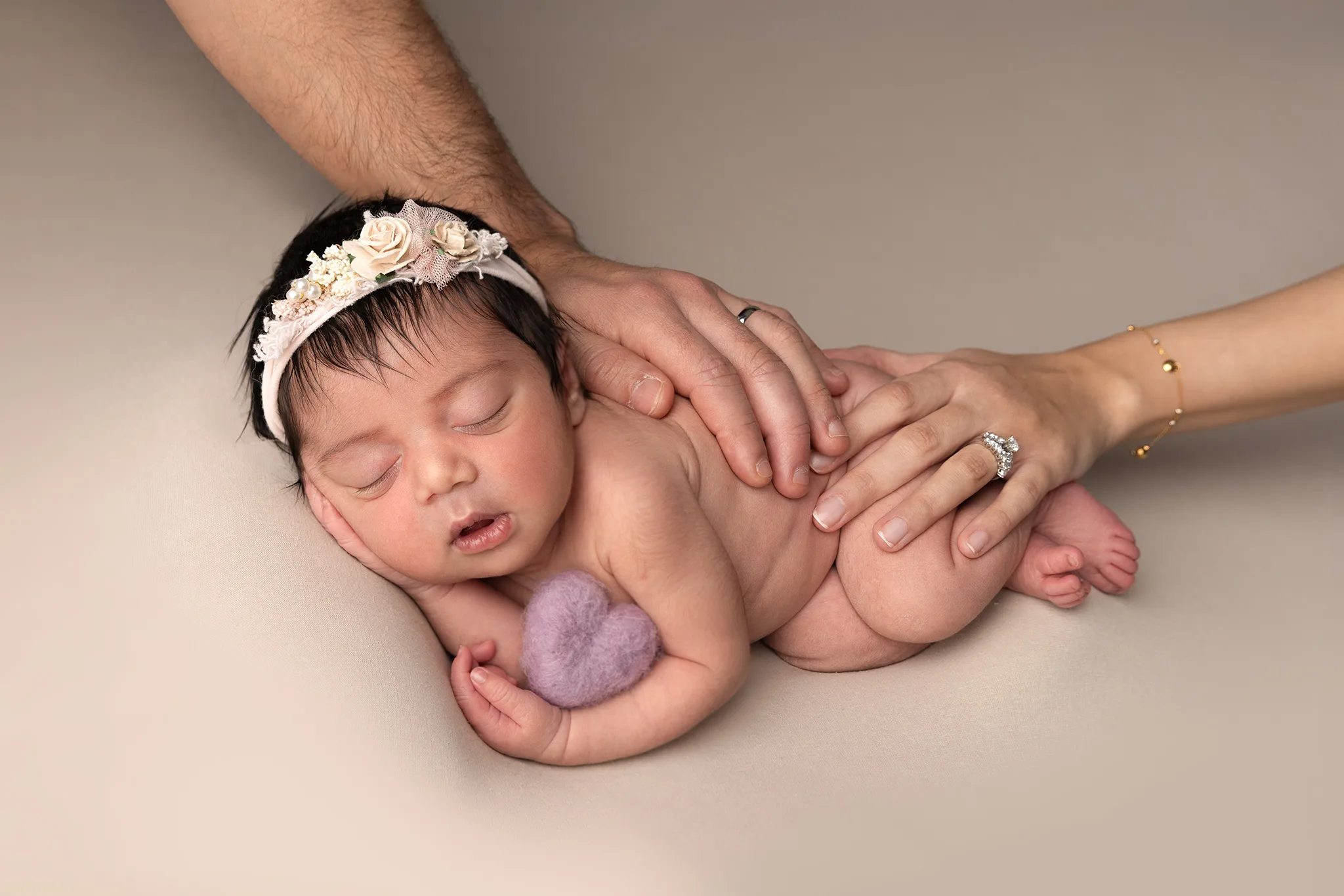 nyfoddfotografering-lund-sovande-bebis-foraldrars-hander En nyfödd bebis sover på beige filt med blommigt pannband, tryggt omgiven av föräldrarnas händer.