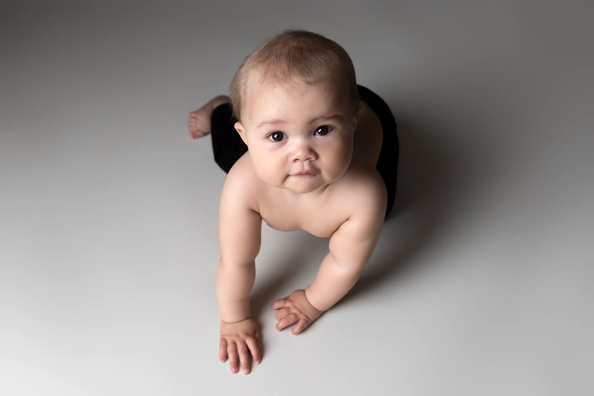 Krypande baby tittar rakt in i kameran mot ljusgrå bakgrund; studiofoto i Lund.