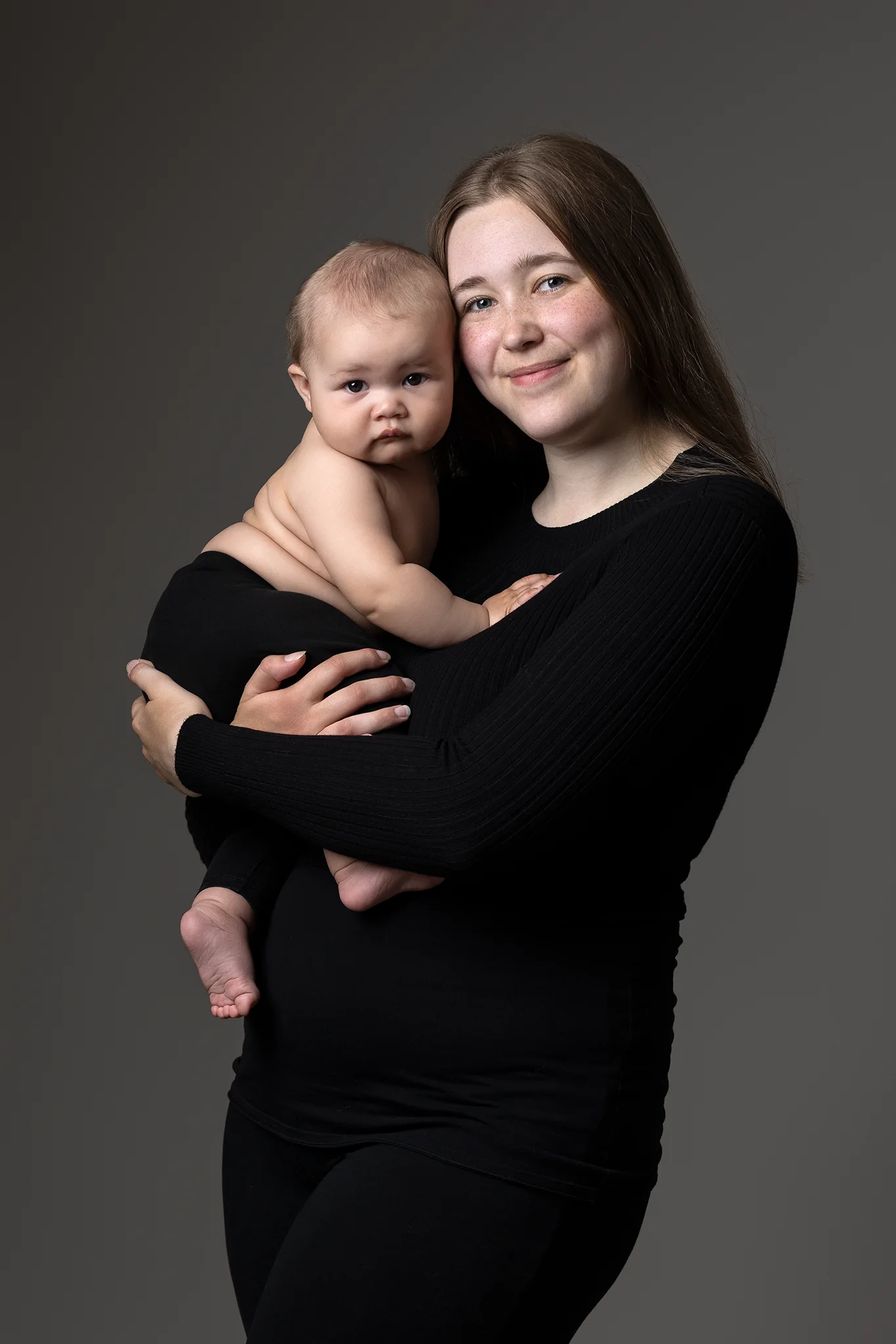 Mamma i svart tröja håller sin nakna baby tätt mot bröstet; porträtt taget i Lund.