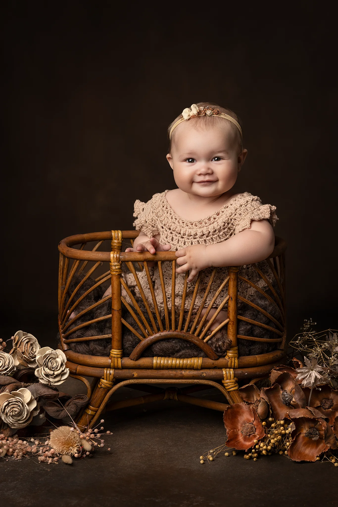 Leende baby står stabilt i rund rottingkorg omgiven av torkade blommor under babyfotografering i Lund.