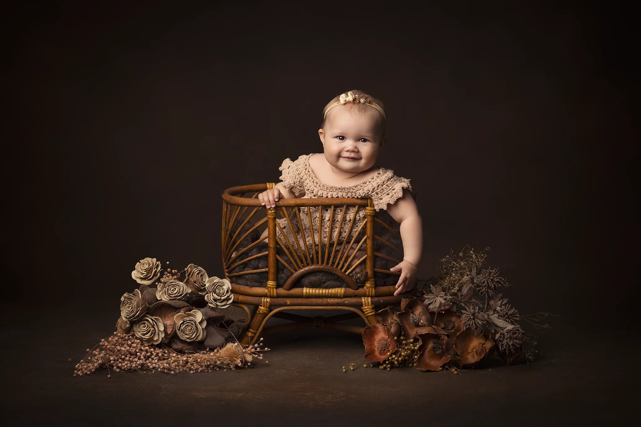 Leende baby står stabilt i rund rottingkorg omgiven av torkade blommor under babyfotografering i Lund.