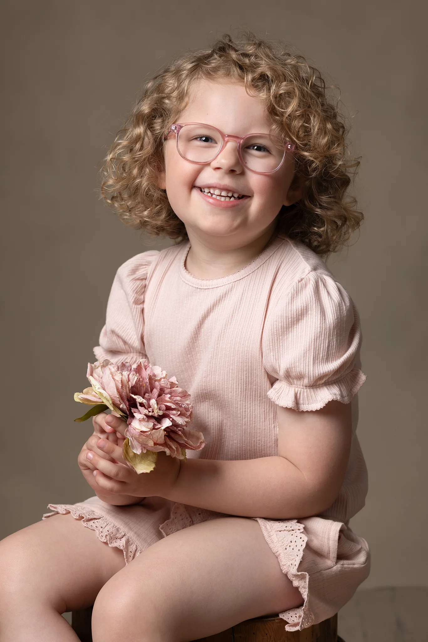 barnfotograf-lund-flicka-med-lockigt-har-rosa-glasogon-blomma Leende liten flicka med lockigt hår, rosa glasögon och blomma fotograferad i studio av barnfotograf i Lund.
