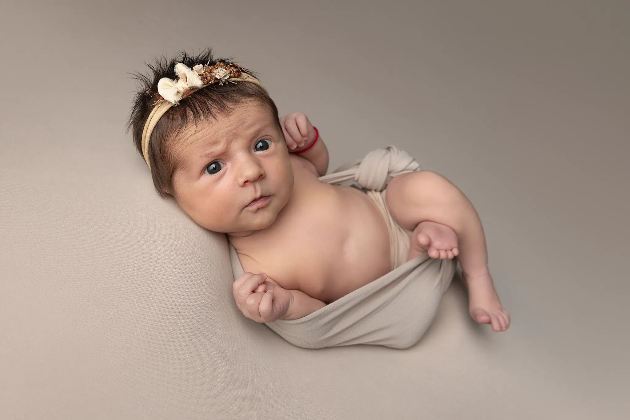 nyfoddfotografering-lund-bebis-harband Vaken nyfödd bebis med blommigt hårband och beige sjal vid nyfö fotografering i Lund.